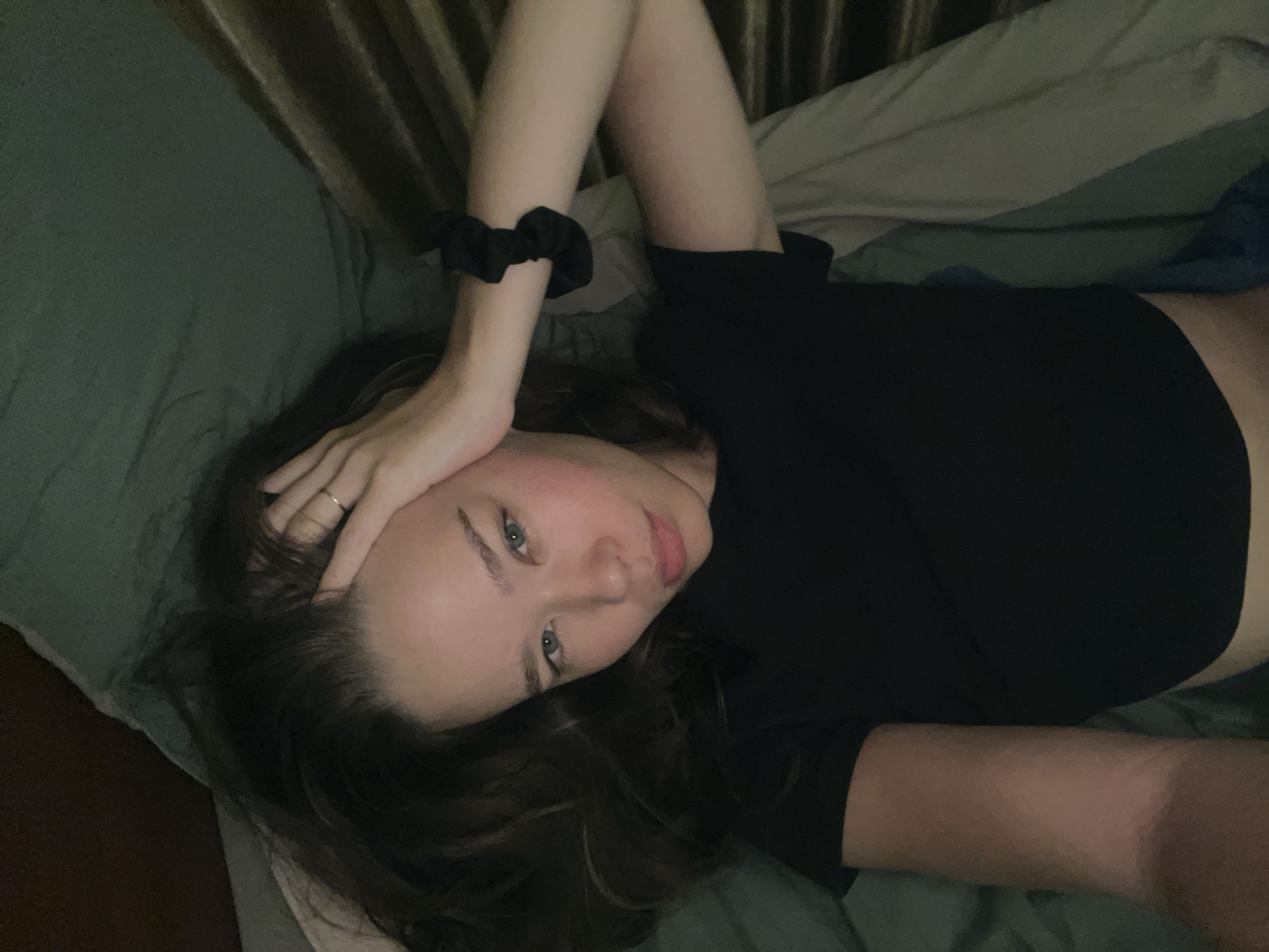 A young woman lies on a bed. She is on her back, with her head turned to the left. Her right arm is raised and her hand is touching her forehead. She has long, dark hair and fair skin. She is wearing a black crop top. A black scrunchie is on her left wrist. The bedding is a mix of green and brown. The location in the city of Nam Nha Trang Ward, Vietnam is not ascertainable from the image.
