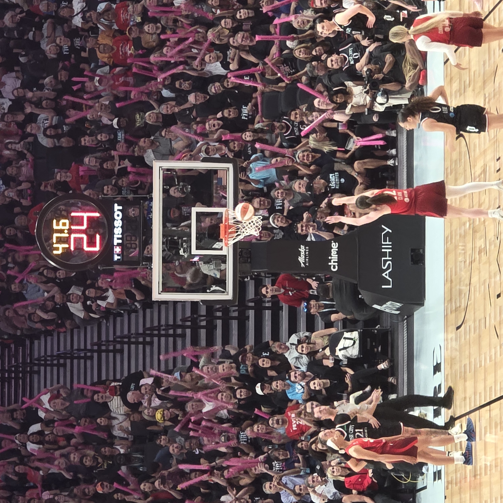 This is a mid-shot photograph taken at the Chiles Center in Portland, Oregon, during a women's basketball game. The court is visible in the foreground with a basketball hoop and a basketball in mid-air, suggesting an active play. Two female athletes in basketball uniforms are present on the court; one in a red uniform with the number 21 on her jersey, and another in a red and black uniform with the number 22. The crowd is visible in the background, seated in tiered stadium seating, many holding pink foam sticks to cheer. A large scoreboard displaying "47.6" and "24" is mounted above the court. Advertisement boards for "Tissot," "Alaska Airlines," and "Lashify" are also visible.