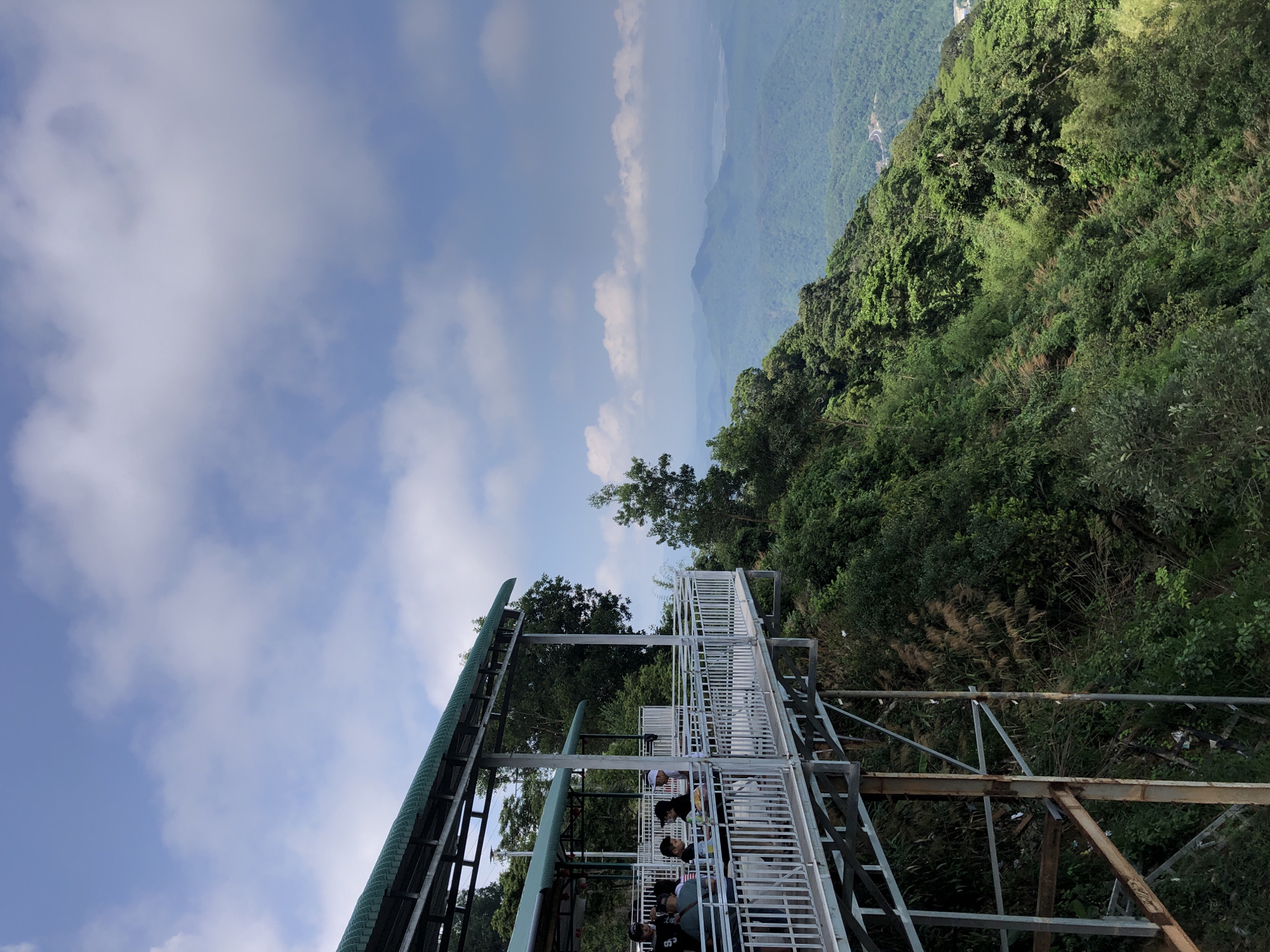 A metal walkway with a green roof extends from the left side of the frame towards the right, elevated above a steep, densely vegetated mountainside. Several people are standing and sitting on the walkway, which is enclosed by white metal railings. The walkway appears to be part of a tourist attraction or observation deck. The background is a panoramic view of lush green mountains and valleys under a partly cloudy blue sky. A body of water can be seen in the distance, nestled among the rolling hills. The scene suggests a scenic overlook in a mountainous region, likely a popular spot for visitors to enjoy the natural landscape. The location is identified as being in Nam Hải, Vietnam.