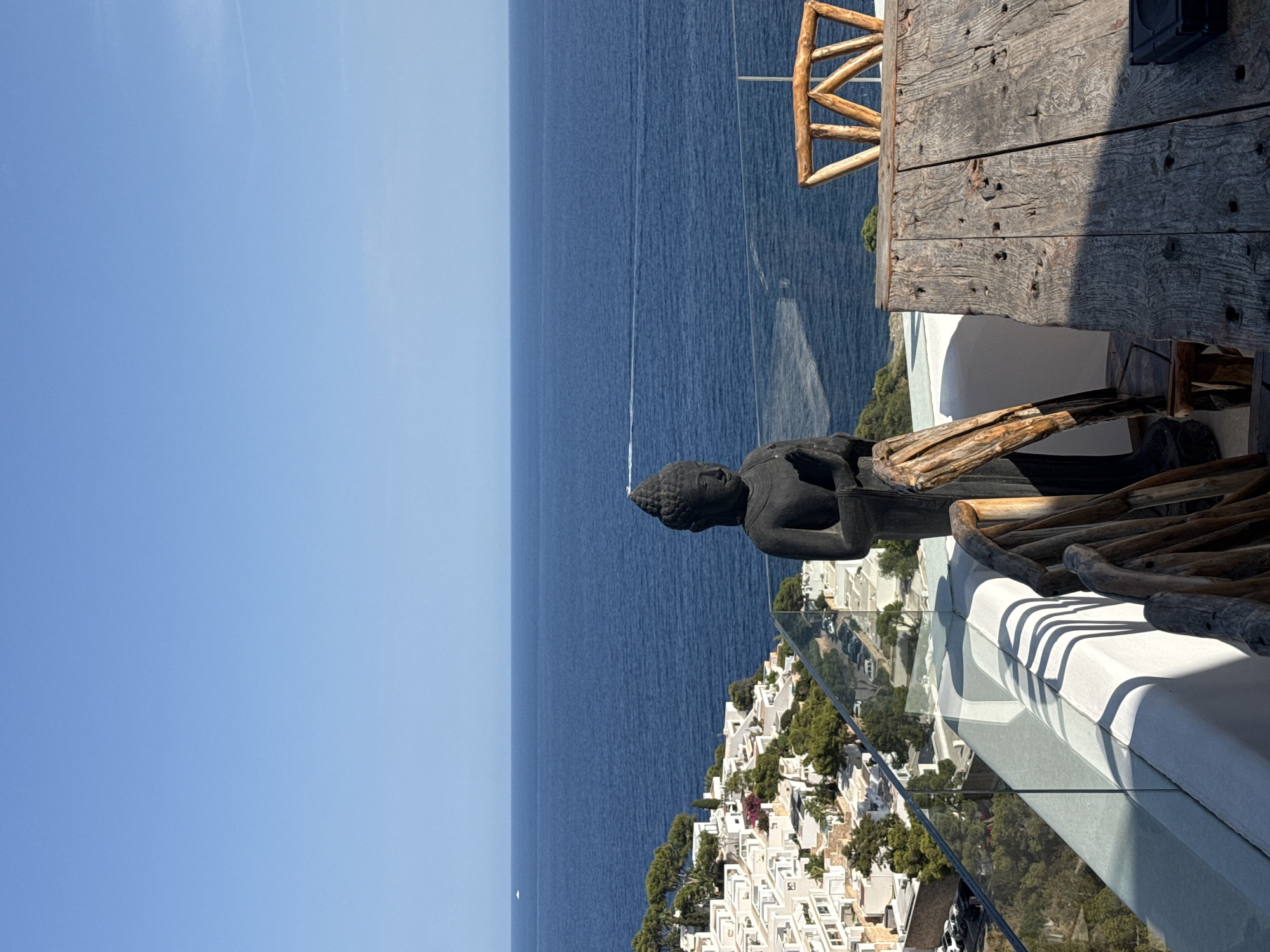 A balustrade with glass panels offers a view of the deep blue Mediterranean Sea. On the balustrade, a dark, weathered statue of Buddha is positioned. In front of the statue, a rustic wooden table and chairs are set up. To the left of the statue, a white building complex with white-tiled roofs cascades down a hillside towards the sea, interspersed with green trees. The sky is a clear, bright blue. The scene suggests a tranquil setting, likely a balcony or terrace overlooking the coastline in Santa Eulària des Riu, Spain.