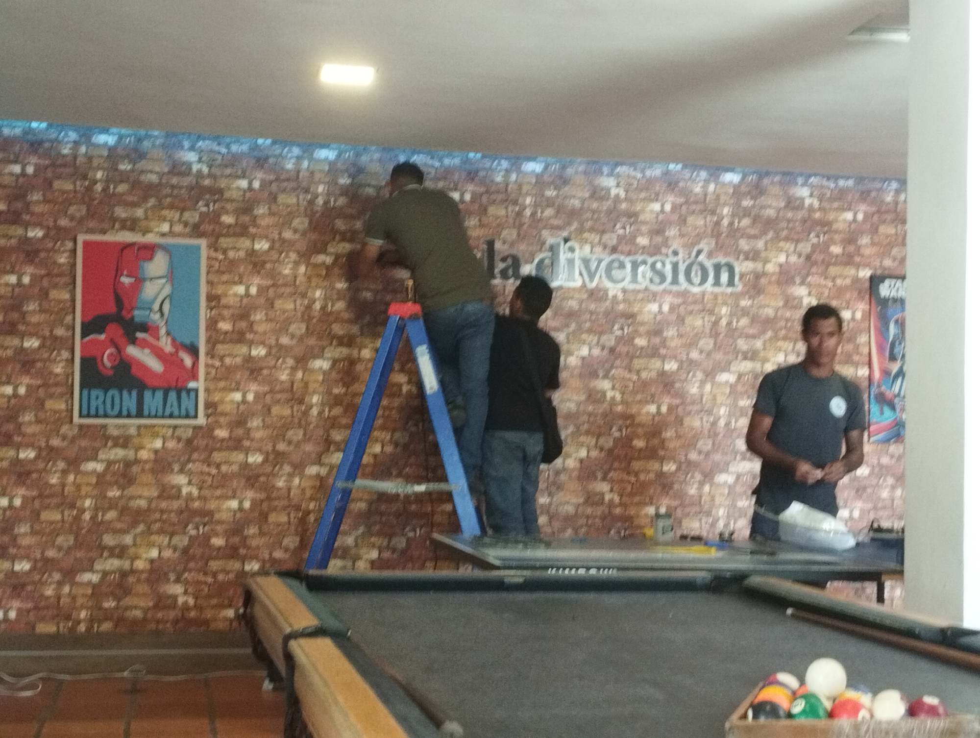 The image shows an indoor scene in Porlamar, Venezuela. Two men are working on a wall with a brick pattern. One man is standing on a blue ladder, appearing to affix something to the wall. A second man is standing next to the ladder, possibly assisting. On the wall is a poster of Iron Man and lettering that reads "la diversión". A third man stands in the background near a table, possibly a pool table, with balls racked. The setting appears to be a recreational or entertainment space.