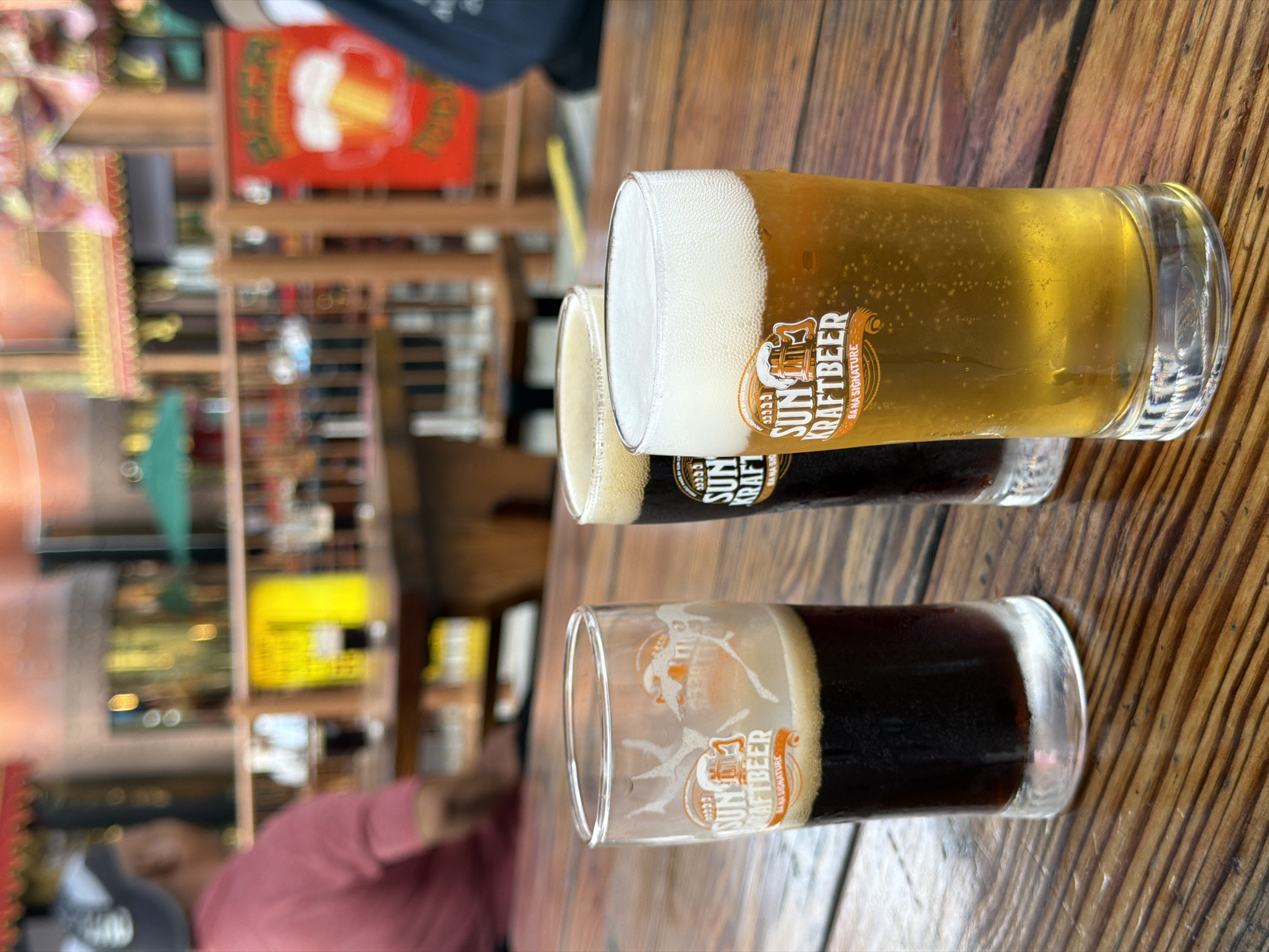 The image shows three small glasses of beer, arranged horizontally on a wooden table. The glasses appear to be tasters, with varying colors and foam levels. A logo for "SUN KRAFTBEER" is visible on each glass. In the blurry background, there is a restaurant setting with shelves of bottles and other items. A person wearing a pink shirt is partially visible. Considering the logo on the glass, it is highly probable that the location of this picture is in Đà Nẵng, Vietnam.