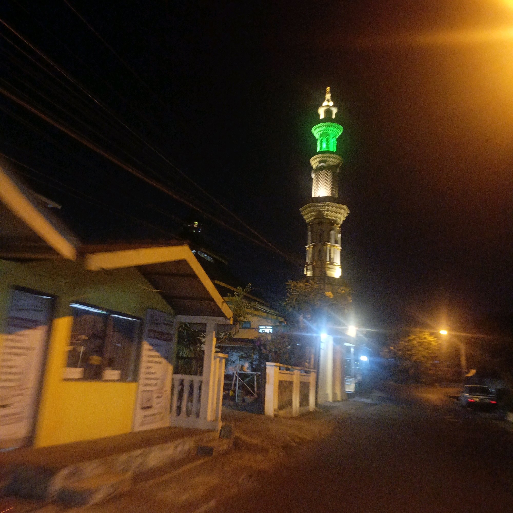 This is a nighttime photograph of a mosque minaret illuminated with green light, located in Salatiga, Indonesia. To the left of the minaret, a yellow building with a covered walkway and a white railing is visible. Power lines are strung across the dark sky. The road in front of the buildings is illuminated by streetlights, and a car is parked on the right side of the road. The overall scene depicts a street in Salatiga at night, with the mosque minaret being the most prominent feature.
