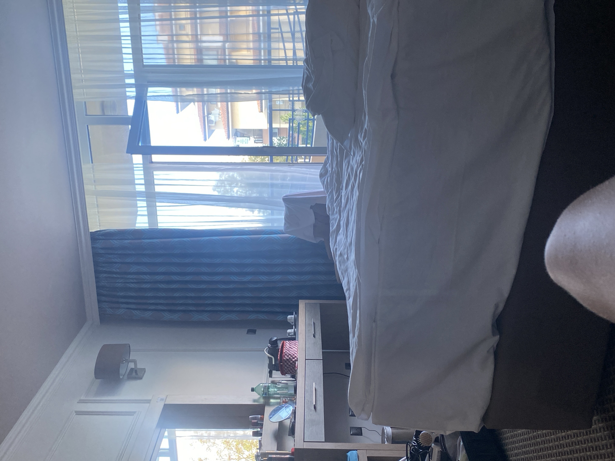 This is an interior shot of a hotel room in Nesebar, Bulgaria. The foreground is dominated by a rumpled bed with white linens. To the left, a wooden dresser with two drawers holds various items, including a red and white checkered box, a coffee machine, bottles, and glasses. A large window with sheer white curtains and blue patterned drapes takes up the background, revealing a balcony and buildings beyond, suggesting a view of an urban or resort area. A wall-mounted light fixture is visible on the left wall.
