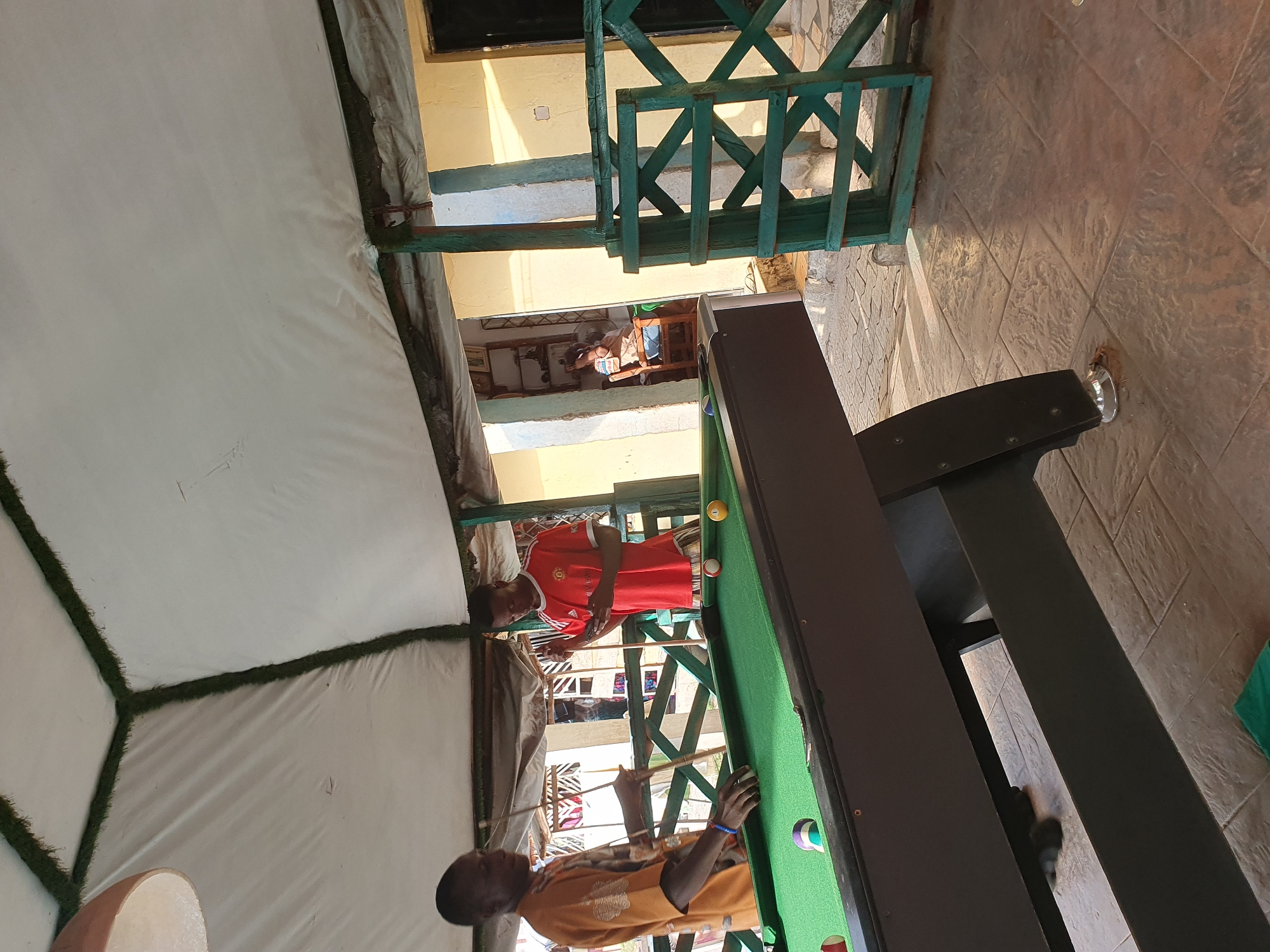 The image captures an outdoor scene, possibly in Lokoja, Nigeria. The foreground features a pool table with green felt, illuminated by overhead lighting. Two young men are actively engaged in playing pool. One player, wearing a red t-shirt, is positioned at one end of the table, seemingly taking a shot. The other player, in an orange t-shirt, is at the opposite end, observing the game. The players appear focused on the game.
The setting is an open-air area, partially covered by a white tent-like structure, with green turf borders visible. Beyond the pool table, there's a green wooden railing, suggesting an elevated platform or balcony. The background reveals a building with cream-colored walls and some architectural details, including windows and what looks like another level. The ground outside the covered area is paved with textured tiles. The lighting suggests it is daytime, with sunlight casting shadows, indicating clear weather. The overall atmosphere seems relaxed and recreational, with the players engrossed in their game. No visible text is present in the image.