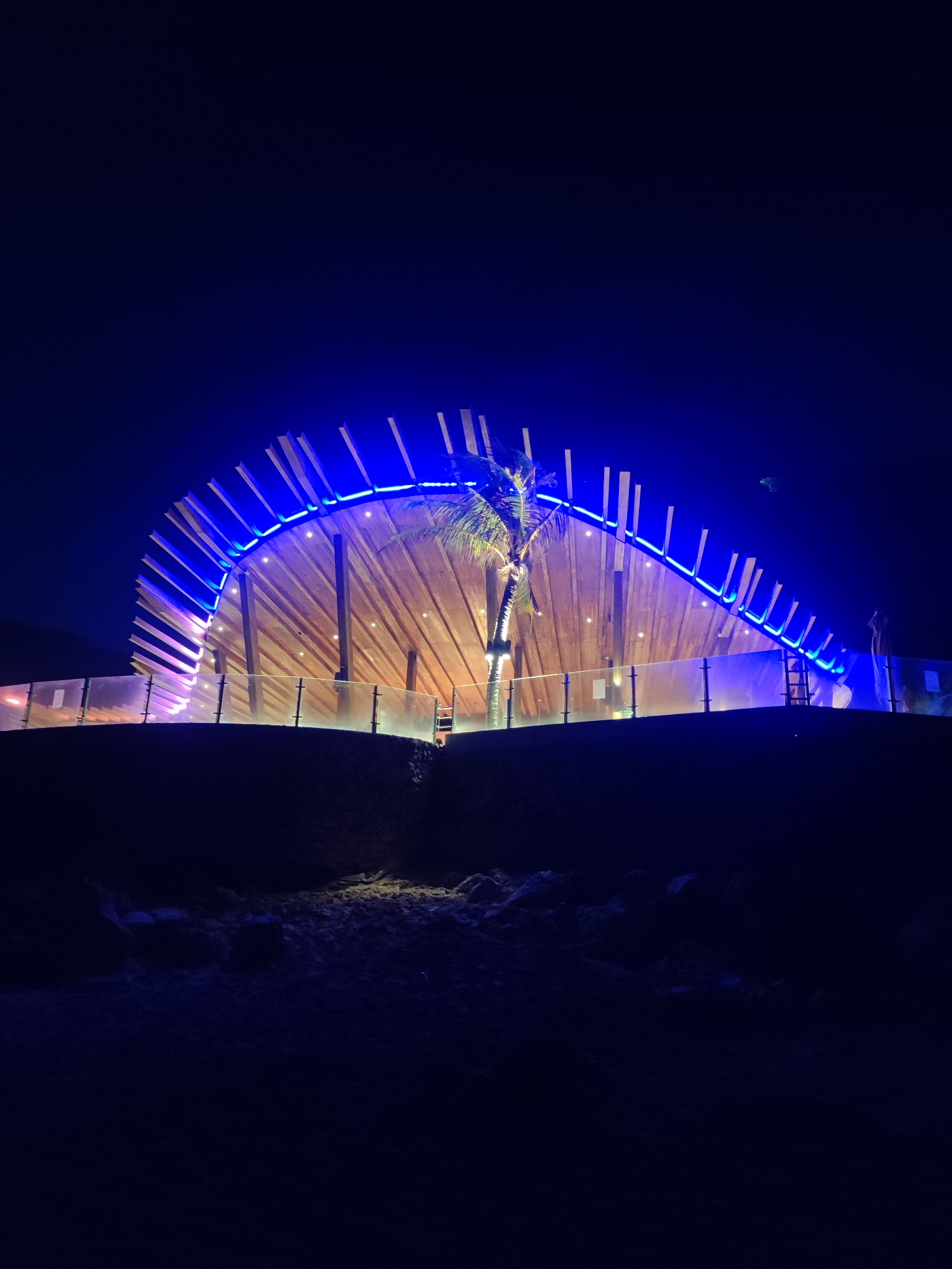 This is a nighttime, low-angle, vertical shot of a modern, semi-circular building with a wooden facade and a curved roof structure, illuminated by bright blue LED lights. The building is situated on a raised embankment behind a transparent glass barrier with metal railings. A palm tree is visible in front of the building. The foreground shows a dark, sandy area with rocks. The scene is located in Kuta Selatan, Indonesia, likely a beachside or resort area, given the presence of palm trees and the architectural style. The overall impression is one of contemporary design integrated with natural elements, highlighted by dramatic nighttime lighting. There are no people visible in the image.
