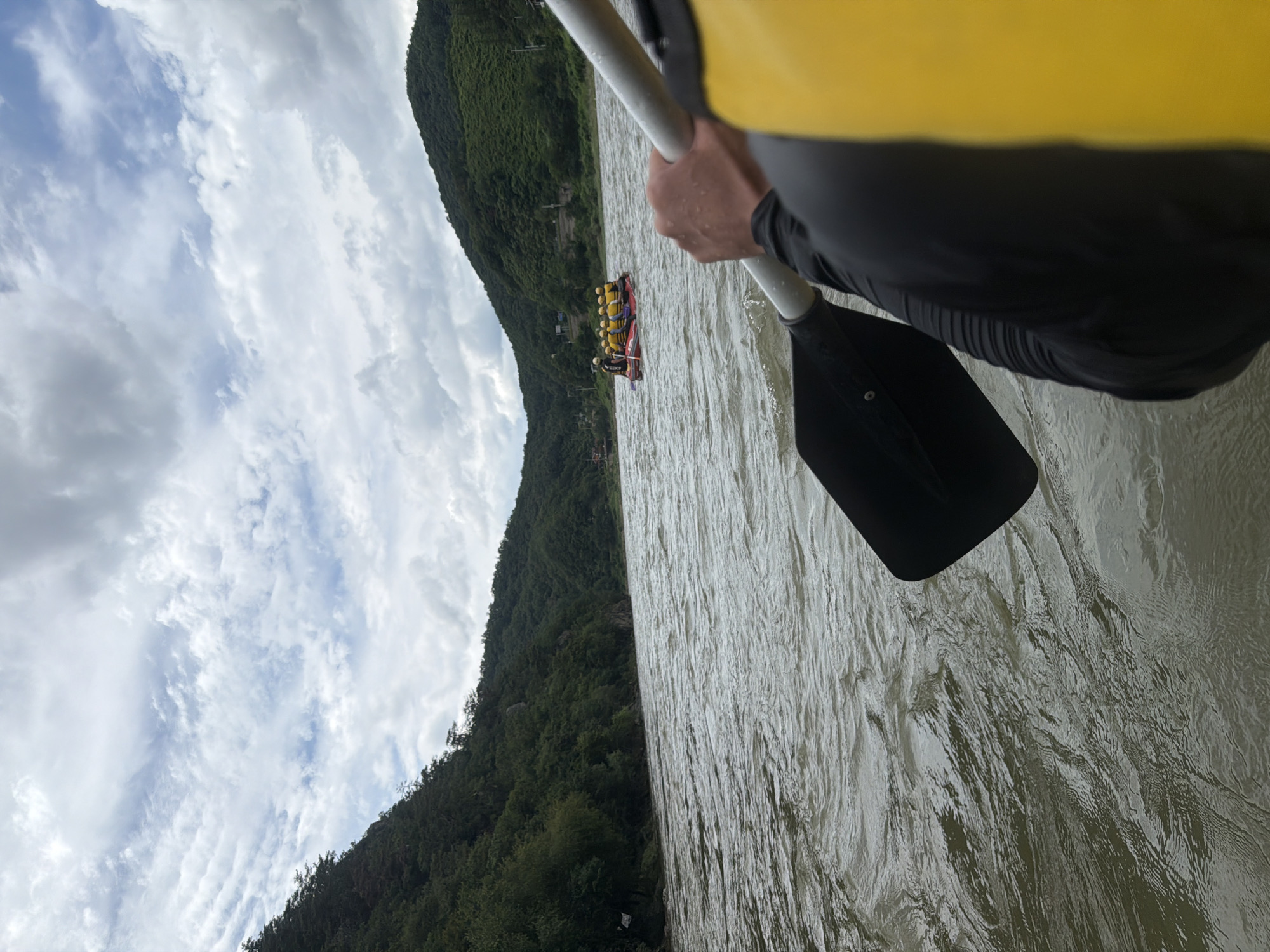 In this perspective shot, a person is holding a paddle, their arm and part of their yellow life vest visible in the foreground. The paddle is poised over the rippling surface of a wide river. In the middle ground, a red raft carrying several individuals wearing yellow life vests is moving down the river. The riverbanks are lined with lush green, tree-covered hills, suggesting a rural or natural setting. The sky above is filled with a mix of dramatic, grey clouds and patches of blue, indicating an overcast or changing weather condition. The scene depicts a rafting activity, likely taking place on the river that flows through Seo-myeon, South Korea, known for its scenic natural landscapes and outdoor recreational opportunities.