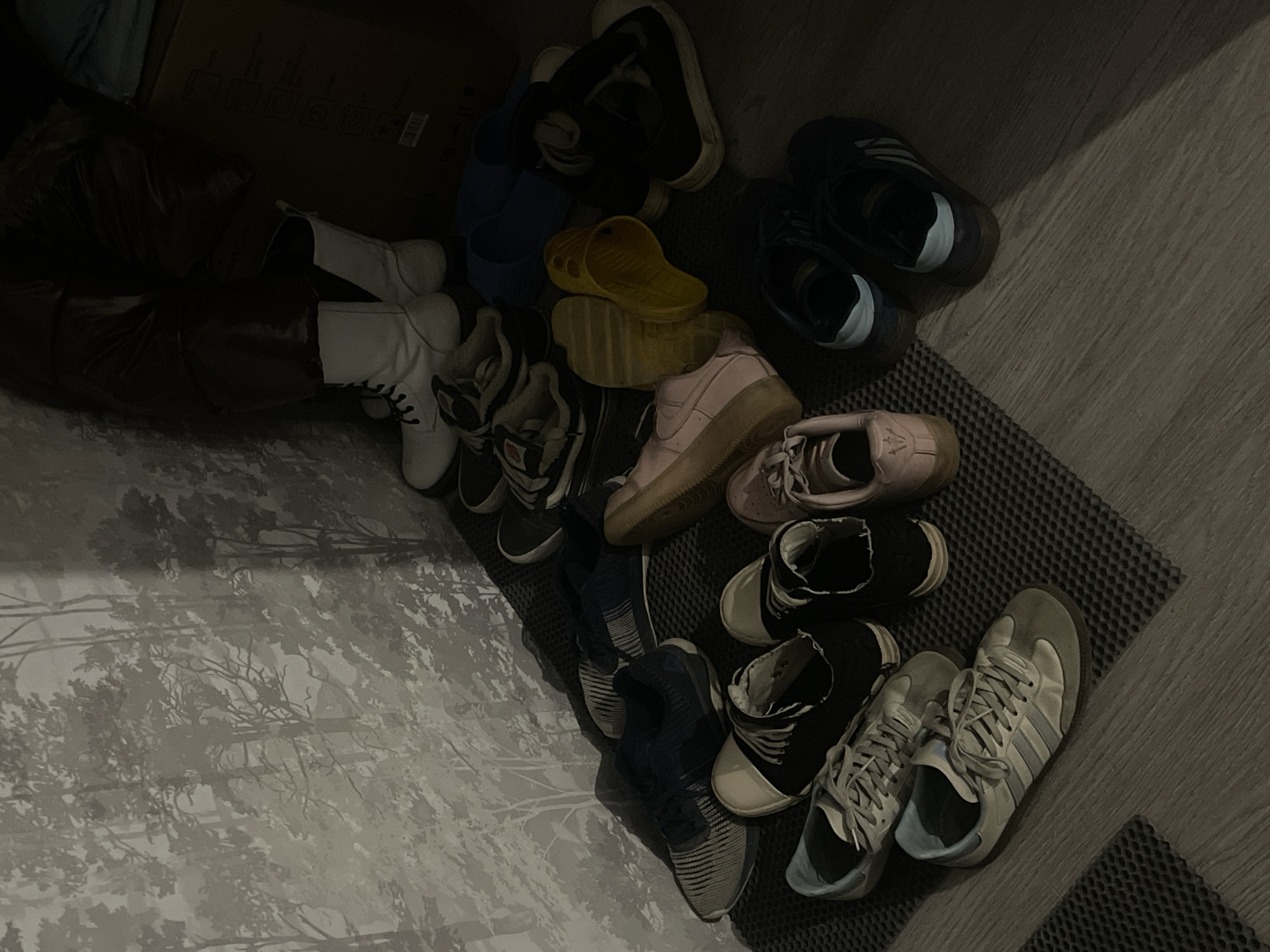 The scene depicts a disorganized collection of footwear clustered on a floor. The footwear includes boots (white and brown), sneakers (various colors), and sandals (yellow). The footwear is situated on a grey mat. The background consists of a patterned wall and a dark cabinet. There is insufficient information to identify the location as Saint Petersburg, Russia. No people, actions, or interactions are visible.