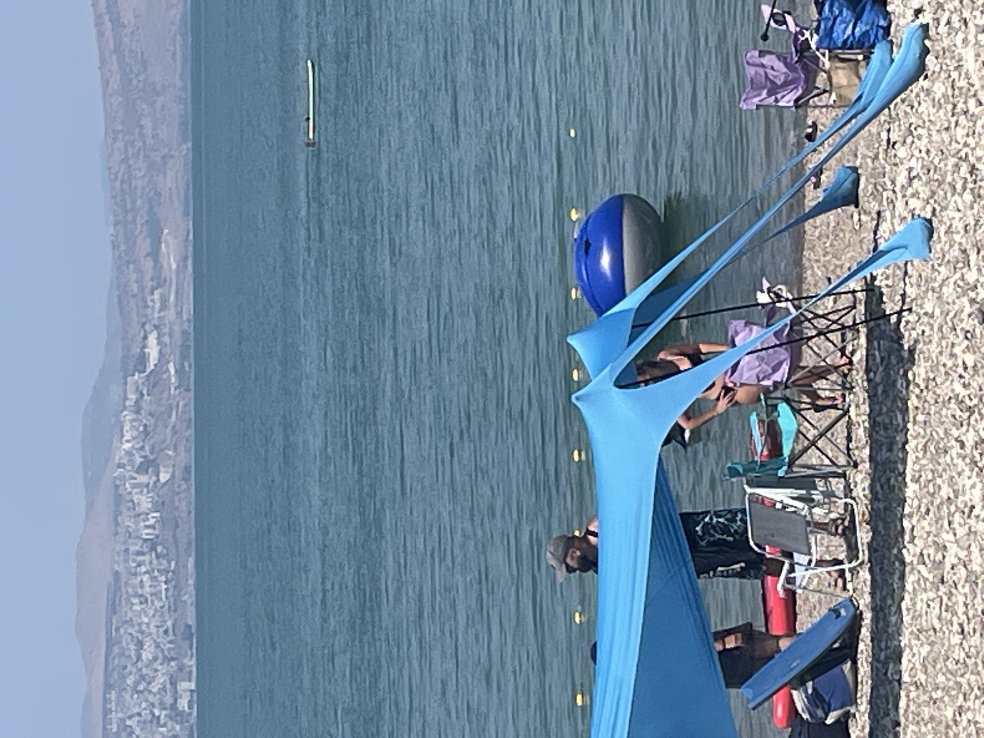 The image depicts a beach scene in the city of Emek HaYarden Regional Council, Israel, with the Sea of Galilee in the background. The foreground is dominated by a blue beach tent, under which several people are gathered. One person, a man wearing a cap and a dark top, stands near the water. To his right, another person, a woman in a bikini, is partially visible.
Scattered on the beach are camping chairs, a red inflatable object, a blue surfboard leaning against a backpack, and a blue inflatable ball floating in the water. The Sea of Galilee stretches out behind them, with a distant cityscape visible on the far shore and mountains beyond. A narrow boat with two people is also visible on the water in the distance. The weather appears to be sunny, with calm water. The beach is covered in pebbles.