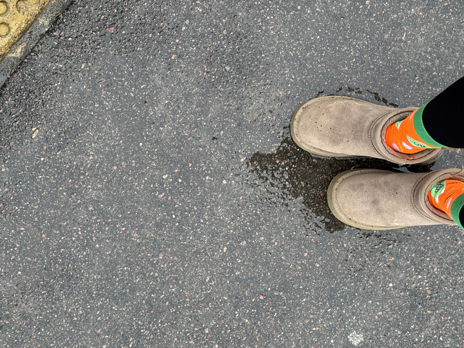 An overhead photograph captures a person standing on a wet, dark gray asphalt surface in Rossosh, Russia. The individual wears light brown, possibly suede, ankle-height boots. Brightly colored socks, primarily orange with green cuffs and white patterns, are visible between the boot tops and the wearer's black lower leg coverings. A small, irregular puddle is present on the asphalt, partially beneath the boots, with additional moisture visible across the surface. An indistinct dark shadow, likely cast by the individual, extends to the left of the boots. In the upper right corner, a segment of yellow tactile paving with circular raised patterns is discernible.