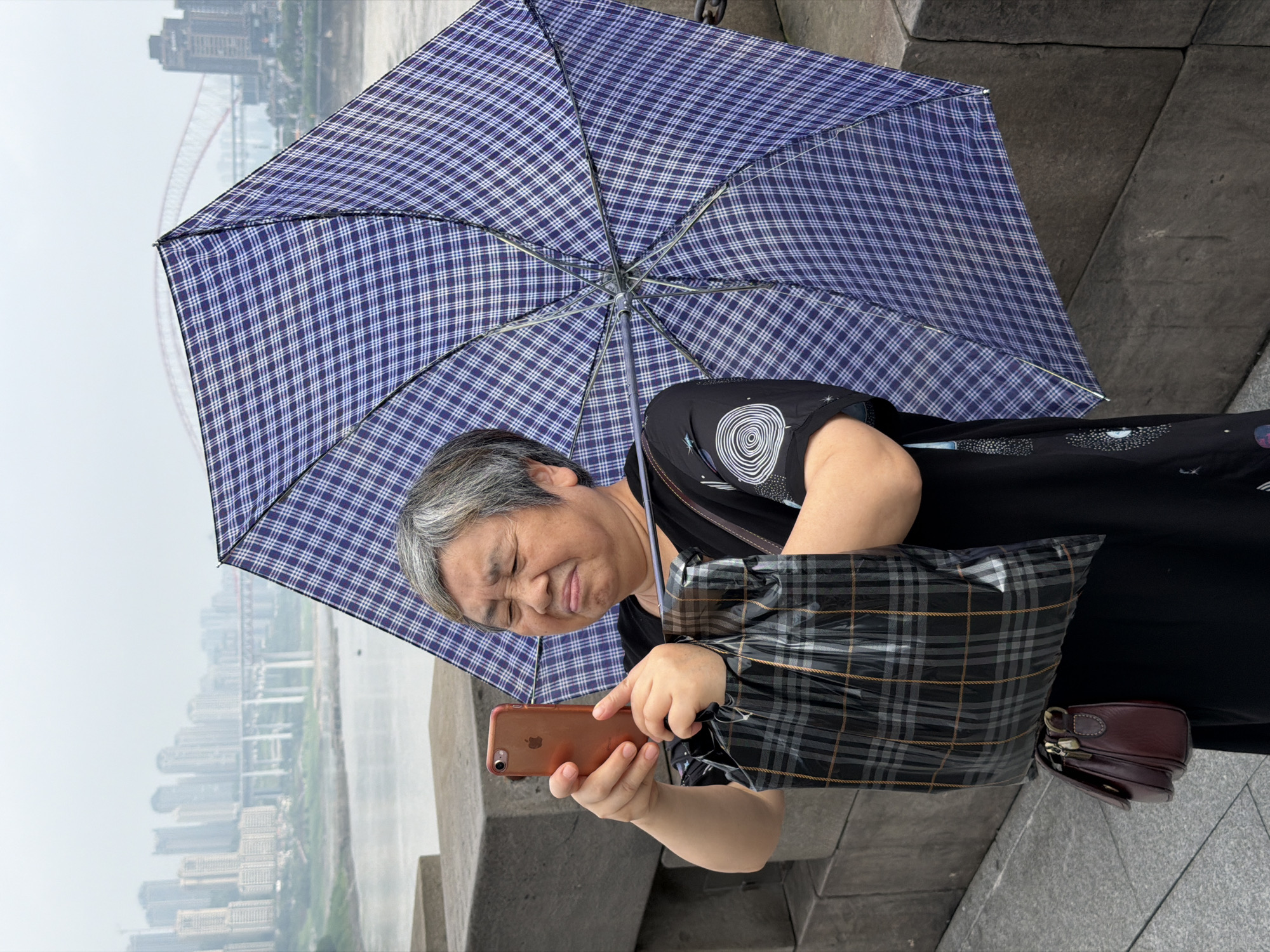 An elderly East Asian woman stands on a raised stone walkway, holding a smartphone in her left hand and a large blue and white plaid umbrella in her right, tilted upwards to her left. Her face is contorted in an expression of discomfort or exertion, with her eyes squeezed shut and her mouth open. She is wearing a black short-sleeved dress with a circular graphic on the left side of the chest. She also carries a large black and grey plaid shopping bag in the crook of her left arm, and a small brown leather purse is visible hanging from her left elbow.
In the background, a hazy cityscape is visible across a body of water. Several tall buildings line the distant shore, and a large red bridge arches across the water. The walkway appears to be part of a waterfront promenade in the Yuzhong District of China, with heavy stone blocks forming a low wall to her right. The sky is overcast, contributing to the diffused light of the scene.