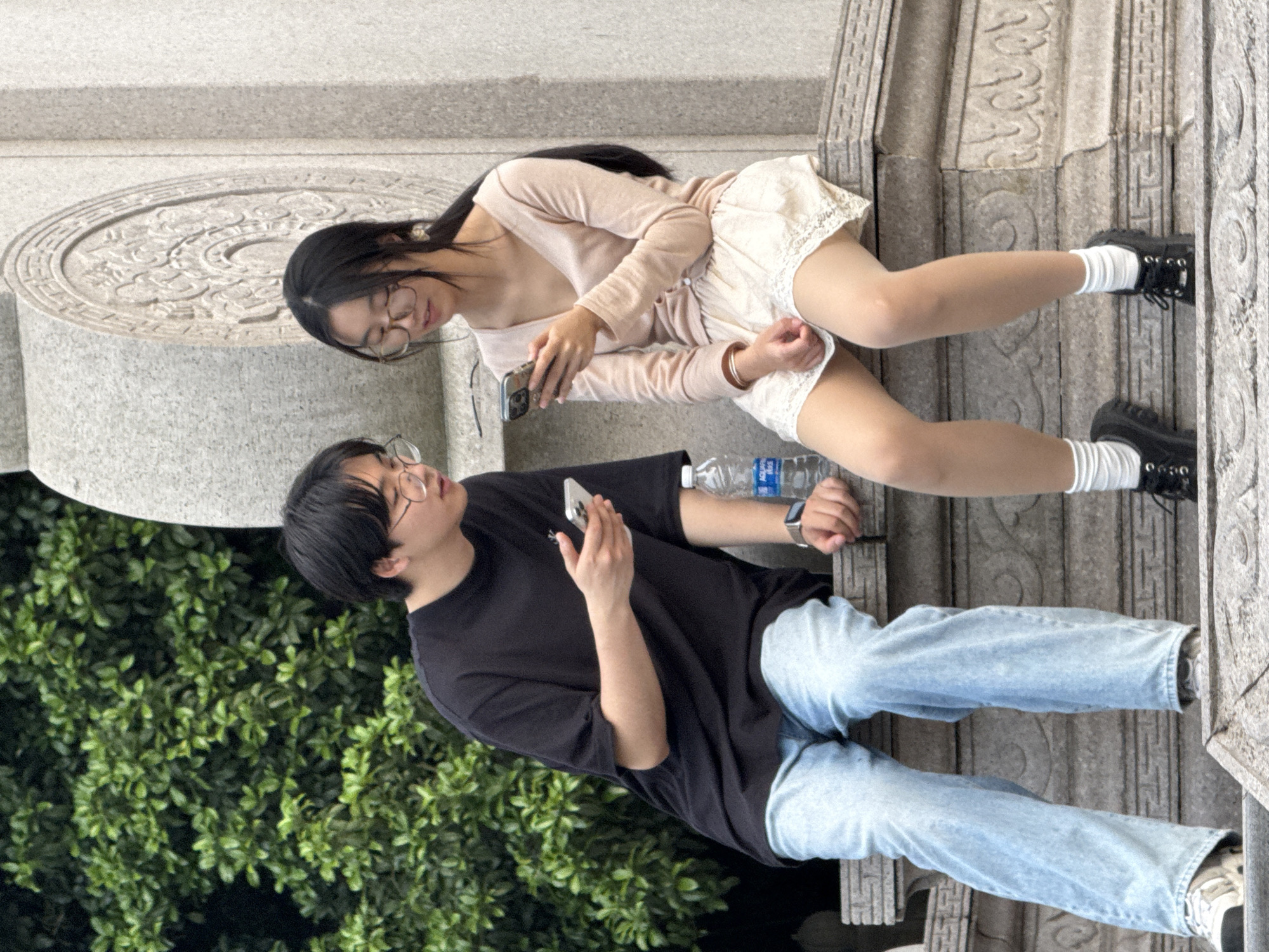Two young adults are sitting on a stone structure. The woman is wearing glasses, a light pink long-sleeved top, and a white skirt with lace trim. She is holding a smartphone. The man is wearing glasses, a black t-shirt, and light blue jeans. He is also holding a smartphone. A water bottle sits between them. They are positioned in front of dense green foliage and what appears to be detailed stonework. The architecture of the stone structure suggests it may be a monument or part of a park in the Liangjiang area.