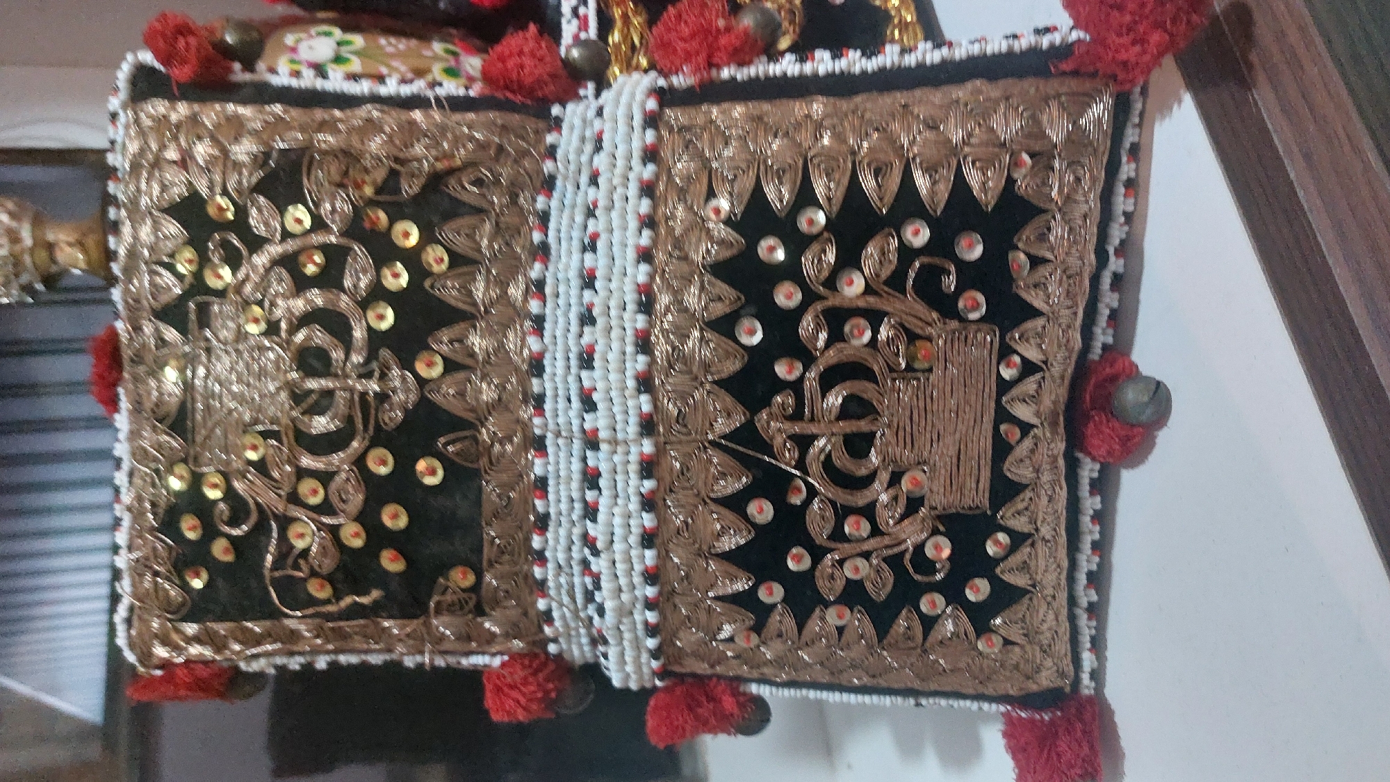A close-up, eye-level shot shows a decorative, possibly ceremonial, pouch or bag. The item is primarily black with intricate gold embroidery and is adorned with small red and gold sequins. A wide band of white, black, and red beads separates the upper and lower sections of the pouch. Red tassels with small metallic bells are attached to the sides and bottom. The background is blurred but appears to be indoors with a wooden surface visible in the foreground. The object is positioned on a white surface. There is no visible indication of location within the City of Medan, Indonesia.