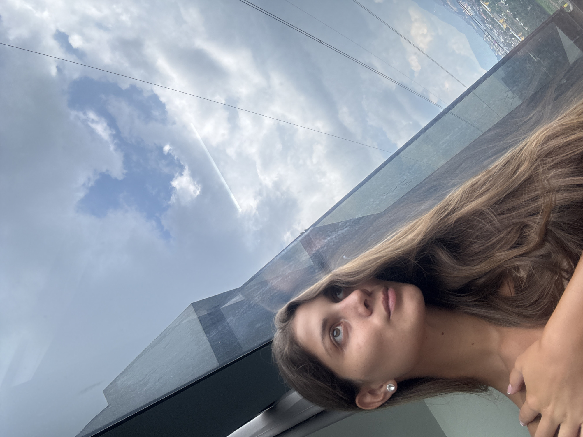The image shows a young woman captured in a thoughtful moment, looking out of a large glass panel. Her head and upper torso are visible, with her long, wavy light brown hair flowing over her shoulders. She wears a simple stud earring, and her gaze is directed slightly upwards and to the left, through the window. Her expression appears serene and contemplative.
She is situated indoors or within an enclosed structure, such as an observation deck or a cable car, indicated by the prominent glass and structural elements visible in the foreground and midground. Thin wires or cables stretch across the sky outside, suggesting an elevated position.
Beyond the glass, the sky is partly cloudy with patches of blue visible. A faint white contrail from an aircraft arcs across the upper sky. In the distant background, a sprawling cityscape or town is discernible, nestled amidst green hills or mountains, characteristic of a high-altitude view. The lighting indicates it is daytime, likely a bright but overcast day.
The scene portrays a tranquil moment of observation from a high vantage point, with the provided context placing this view in Kuala Lumpur, Malaysia. There is no visible text in the image.