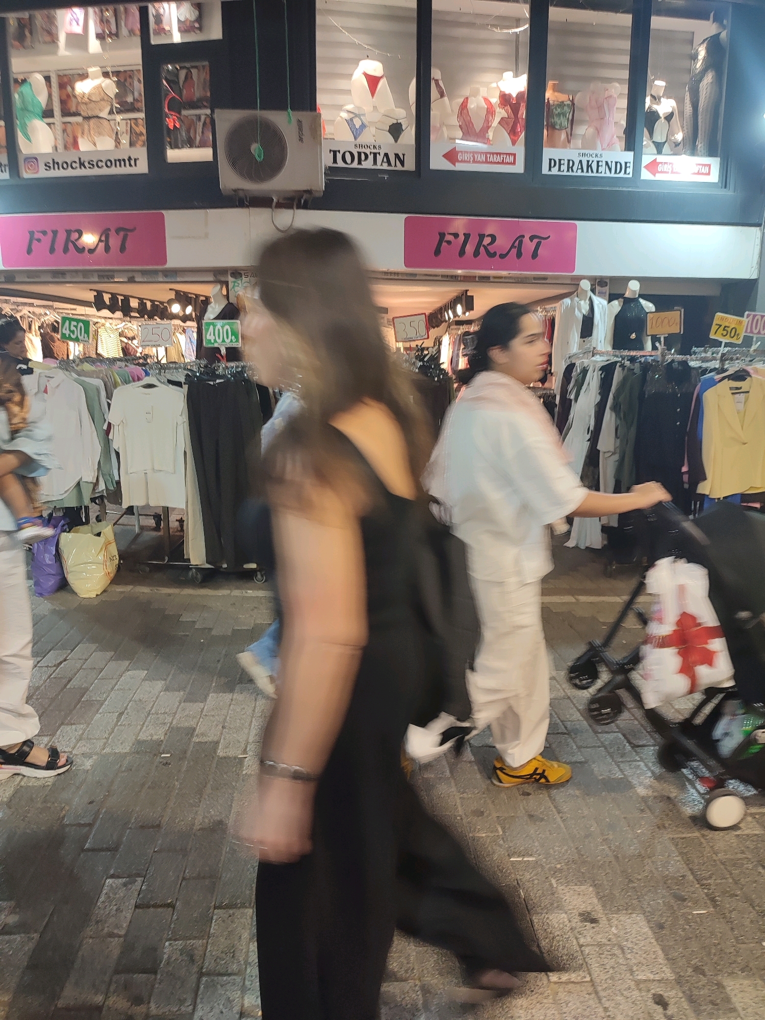 The image depicts a street scene in Kadıköy, Turkey, likely on a pedestrian walkway lined with shops. In the foreground, a person in a black dress is blurred due to motion, with their arm extended and wearing a bracelet. To their right, another person wearing a white outfit and yellow shoes is pushing a stroller with a white bag adorned with a red ribbon. Behind them, a clothing store is visible, with racks of apparel displayed outside. Signs indicate prices in Turkish Lira, such as "450Ł," "250Ł," and "750Ł." The shop windows showcase mannequins dressed in lingerie and clothing. The street is paved with rectangular stones. The overall impression is a busy commercial area at what appears to be evening or night, with artificial lighting illuminating the scene.