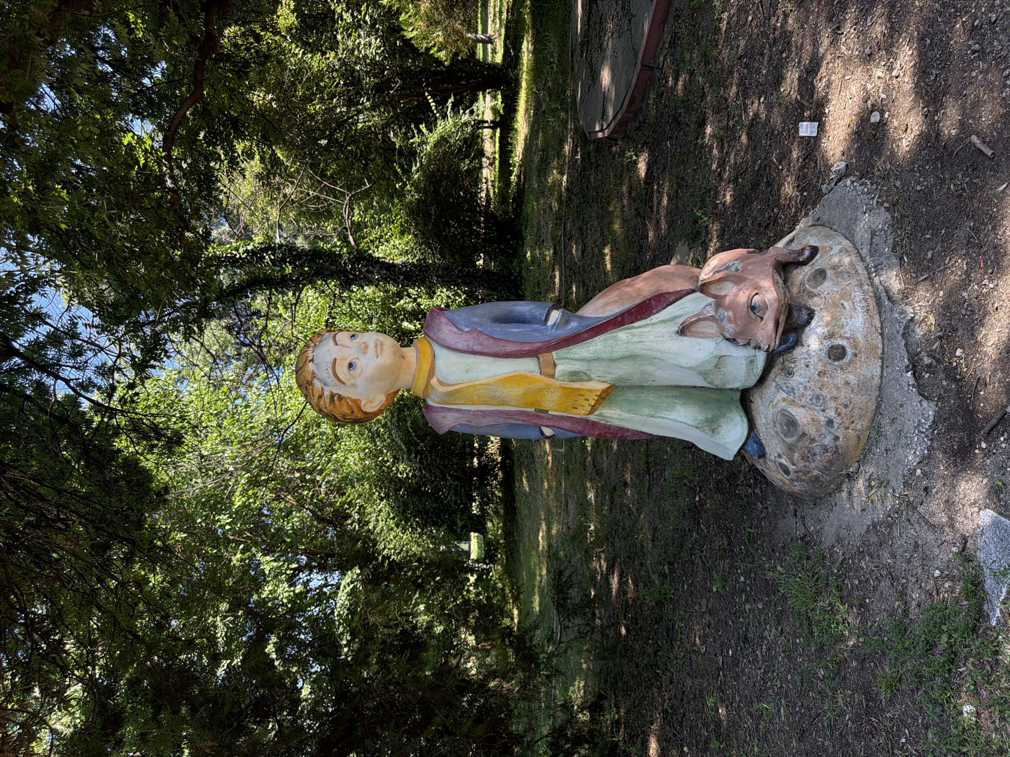 The image depicts a statue of the Little Prince with a fox at its base, located in the City Park of Pazardzhik, Bulgaria. The statue is positioned on a rounded, planet-like pedestal with circular indentations. The Little Prince is rendered in a standing posture, wearing a yellow scarf, a blue coat over a purple garment, and a pale green skirt. His gaze is directed upwards and to the left. The fox, in a resting position, is positioned to the right of the Little Prince's legs. The statue is situated on a dirt and grass-covered ground, partially shaded by mature trees with dense foliage overhead. In the background, the park landscape features more trees, bushes, and a worn, circular concrete structure with a red border. The overall scene is a sunny day with dappled sunlight filtering through the leaves.