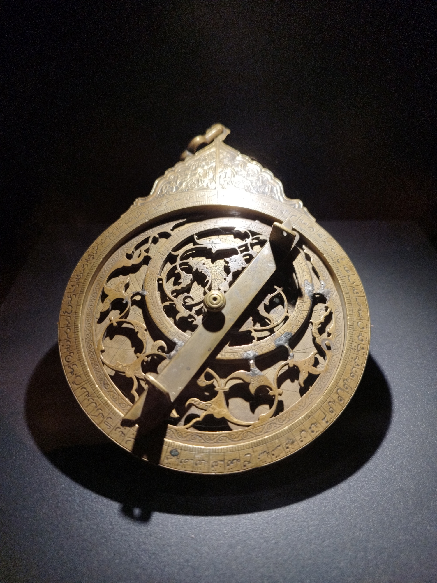 An antique, ornate brass astrolabe is displayed on a dark, textured surface. The astrolabe is circular with a flat, disc-like base adorned with intricate carvings and Arabic inscriptions around its perimeter. A movable pointer, or alidade, is pivoted at the center, extending across the face of the instrument. The alidade is also made of brass and appears to have notches or markings on its surface. The astrolabe's design includes a detailed rete, a latticework overlay depicting celestial star points. The background is dark and appears to be a void or a dimly lit display case, with a spotlight illuminating the astrolabe from above, creating a strong shadow. The overall impression is of a museum or exhibition setting, highlighting a historical artifact.