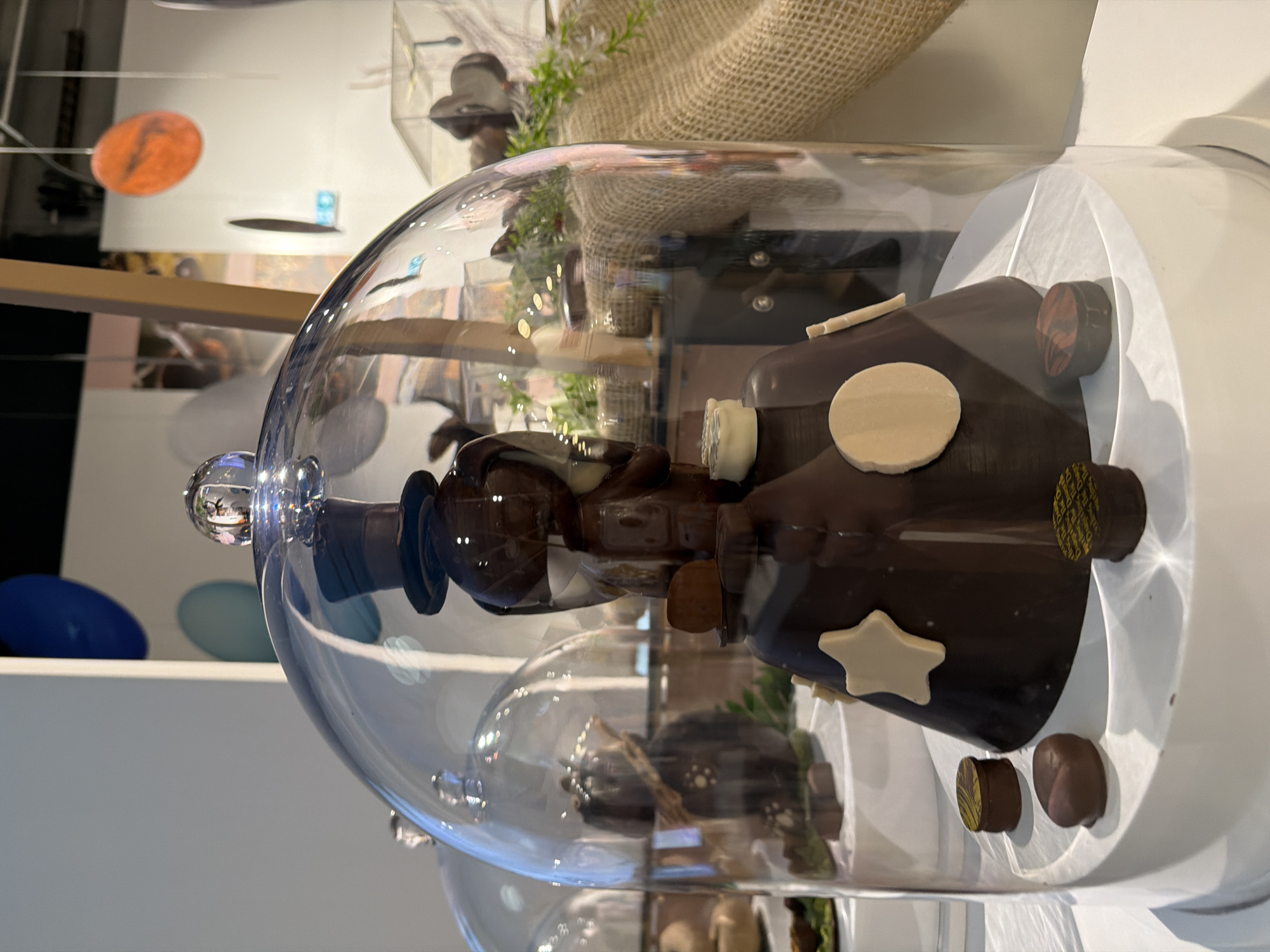The image shows a chocolate sculpture under a glass dome. The sculpture is predominantly dark brown and decorated with circular and star-shaped white chocolate pieces and smaller chocolate roundels. The sculpture rests on a white, cylindrical base. The background reveals a partial view of a building interior, showcasing various decorative items and architectural details. The image was taken at Illkirch-Graffenstaden, France.