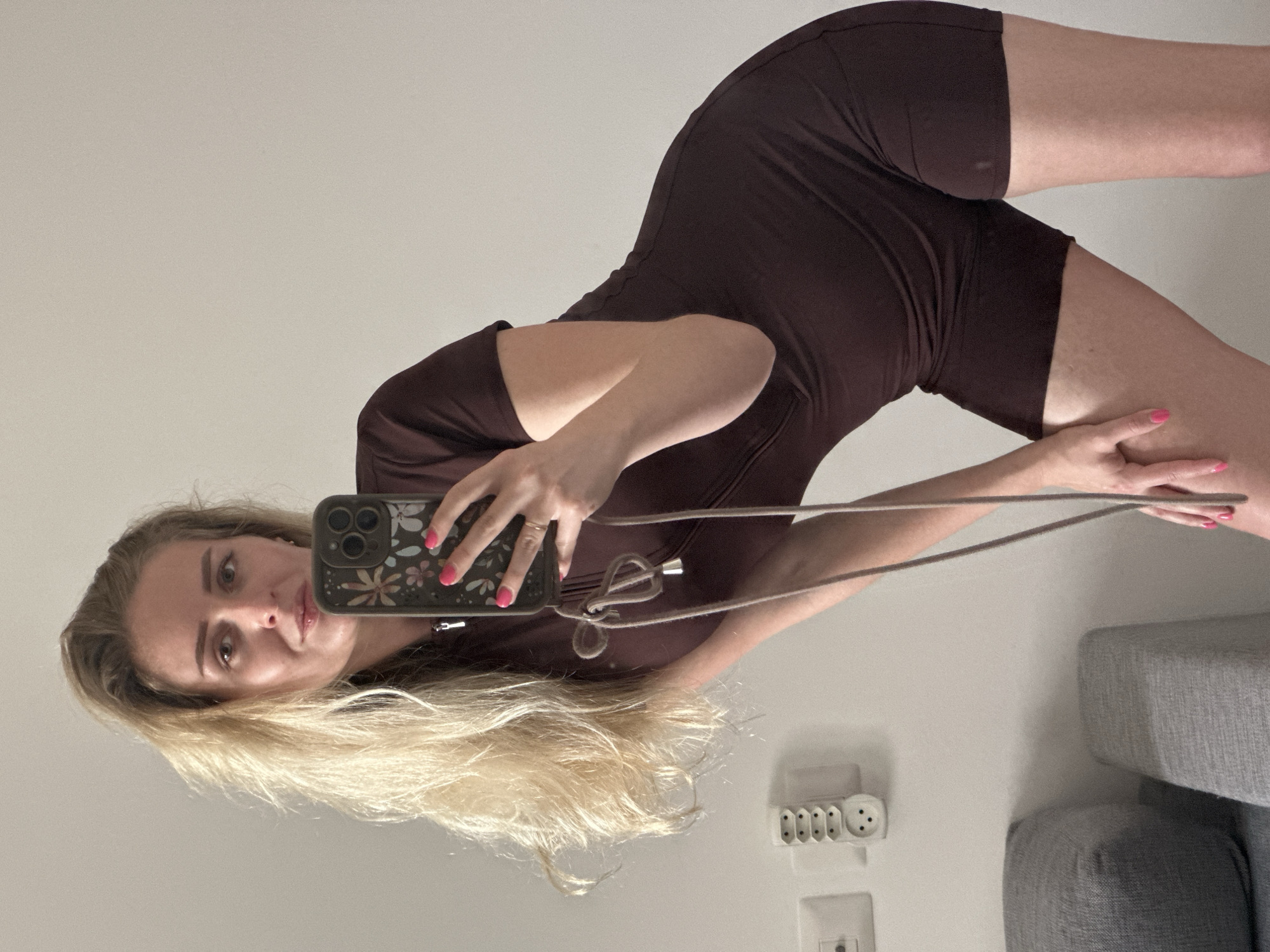 This image is a mirror selfie featuring a young woman indoors, likely within a home in Petah Tikva, Israel. The woman, with long blonde hair, is shown in a dynamic pose, looking directly into the camera lens of her smartphone. She is wearing a dark brown or maroon, short-sleeved, short-legged romper or jumpsuit. Her left hand holds the phone, which is encased in a dark cover adorned with a subtle floral pattern, and a light-colored lanyard or strap is attached to it. Her right hand, sporting pink nail polish and a ring on the ring finger, rests on her right thigh.
The background is a plain, light-colored wall. On the lower right side of the wall, a white electrical power strip with multiple European-style sockets is visible, positioned above a white square wall panel that appears to be a light switch or another outlet. To the far right, a grey upholstered section of furniture, possibly an armrest or backrest of a sofa or chair, is partially seen. The entire image is presented rotated 90 degrees counter-clockwise from an upright perspective. No explicit text is visible in the frame, but the style of electrical outlets offers a subtle geographical clue consistent with the provided location context.