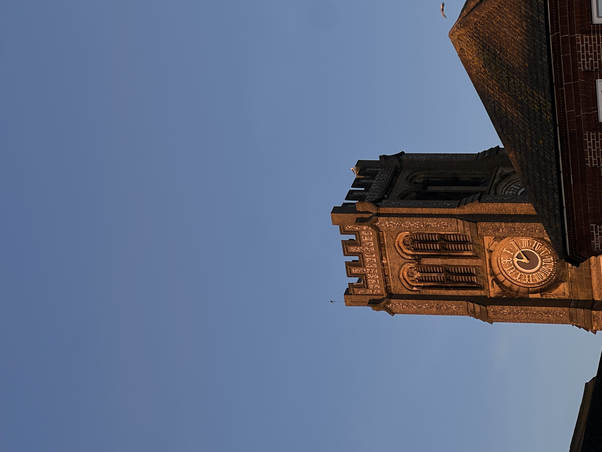 The image depicts the clock tower of St. Mary Magdalene Church in Gorleston-on-Sea, United Kingdom, viewed from a low angle. The sky is visible on the left side. The brickwork of the tower, and the clock face showing 04:38, are illuminated by the sun. A bird flies in the sky. A portion of an adjacent building is also visible.