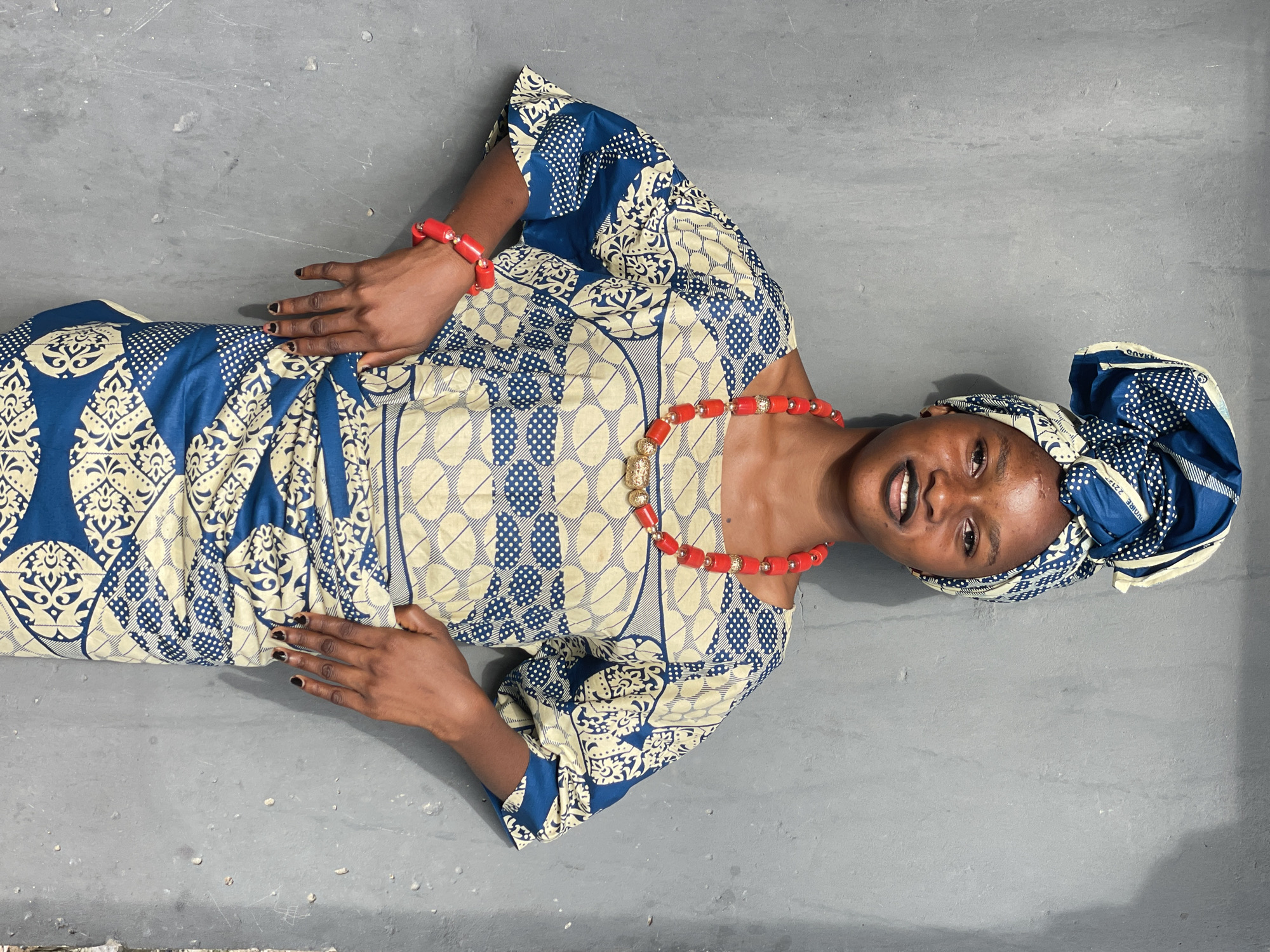 A young woman lies on a flat gray surface, likely a floor or pavement, posing for the camera. She is wearing a traditional patterned blue and white dress and matching headwrap. Her attire is complemented by a red beaded necklace and bracelet. She appears to be in an indoor or covered space due to the even lighting. The context of Badagry, Nigeria, is not explicitly evident from the image alone but can be inferred from the style of clothing and accessories. There is no immediate way to confirm that this image was taken there from the image itself.