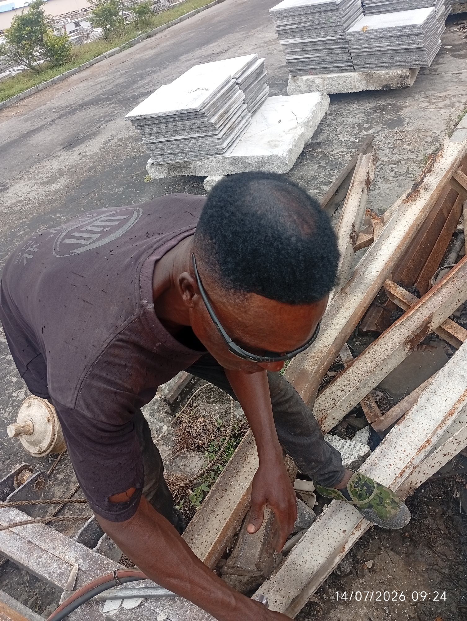 A man, appearing to be of African descent, is seen from a high angle, working outdoors in what seems to be an industrial or construction area. He is wearing a dark t-shirt with a faded circular logo on the back and dark trousers, one leg of which is torn near the knee. He also wears camouflage-patterned slip-on shoes and dark-rimmed safety glasses. The man is bent over, his hands engaged with some metal structures and equipment on the ground.
The environment is paved with asphalt or concrete, and stacks of large, flat granite slabs are visible in the background, suggesting a stone yard or a processing facility. Some of the granite is neatly stacked, while other pieces are scattered or broken. The ground around the work area is uneven and contains debris, including rusty metal beams, wires, and dry vegetation. In the far background, there are some green trees and buildings. The lighting suggests it is daytime. A timestamp in the lower right corner reads "14/07/2026 09:24". The location is identified as Bana Stream, Nigeria.