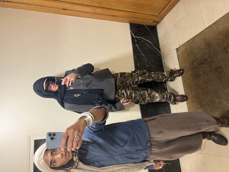 Two individuals pose for a photo in a hallway. One person wears a gray blazer, camouflage pants, brown shoes, a black headscarf, and black-framed glasses. The other is dressed in a blue jacket, tan pants, brown boots, and a beige head covering. Both are holding phones, presumably taking the picture in a mirror, as reflected. The hallway features a light-colored wall, a dark marble border, and a brown rug.