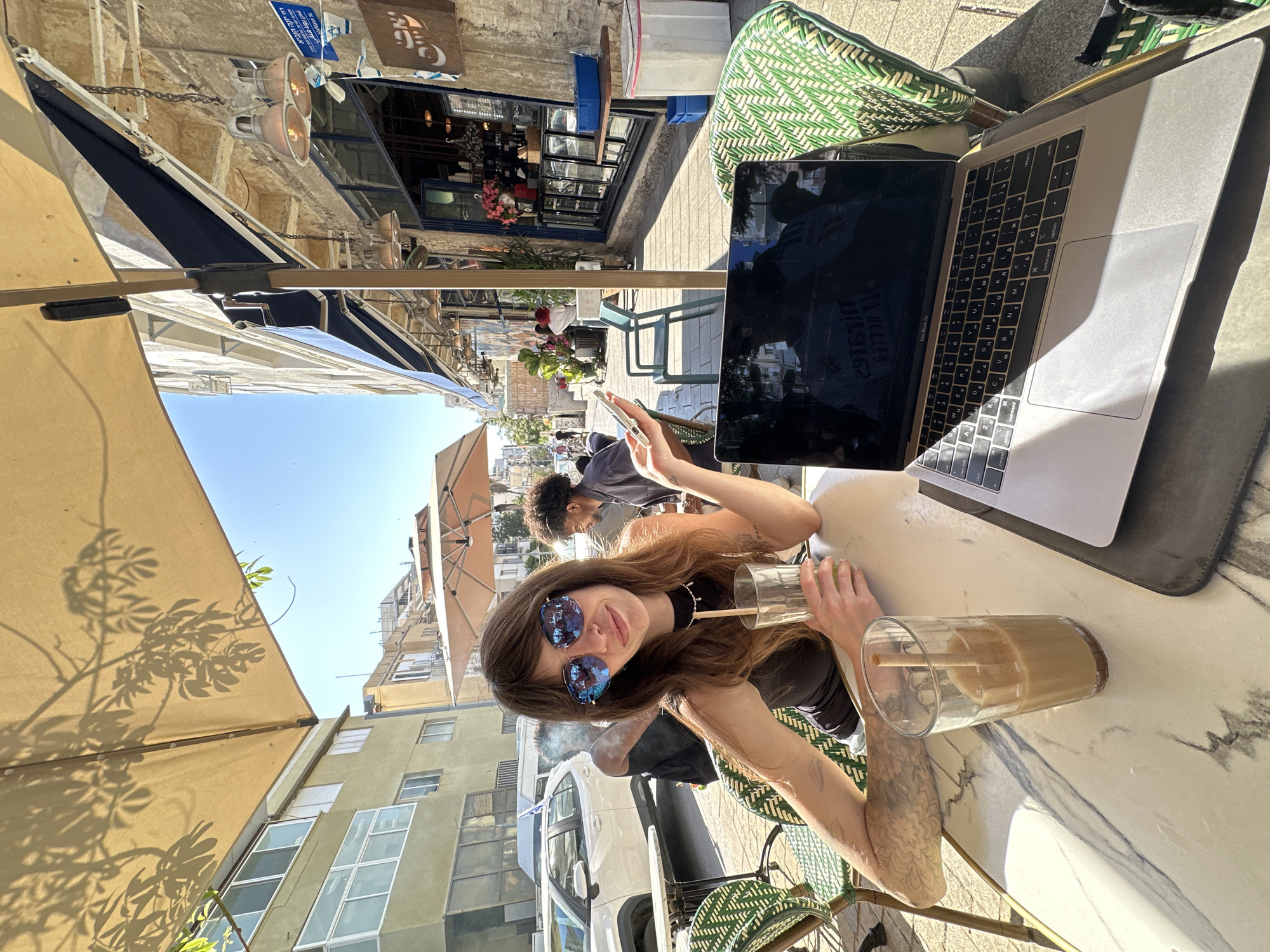 This image captures an outdoor cafe scene, likely in Tel-Aviv, Israel, under bright daylight. The main subject is a woman seated at a marble-patterned table, looking towards the camera. She has long brown hair, wears a dark top, and reflective blue sunglasses. A tattoo is visible on her left forearm. She holds a clear glass with a straw, which contains a beverage. Another glass with an iced drink sits on the table.
An open silver MacBook Air laptop is positioned to her right, displaying a black screen with reflections of the surroundings. The laptop keyboard features both English and Hebrew characters, indicating its use in an Israeli context. Beside the laptop, a green patterned chair and a grey cushion are visible.
In the background, a bustling street scene unfolds. Buildings with various architectural styles, some featuring balconies and windows, line the street. Several umbrellas provide shade over other tables. A blue sign with Hebrew text, possibly including "Tel Aviv," hangs above a storefront, further confirming the location. Another shop front with a brown, stylized logo is also visible. A man with curly hair is seated behind the main subject, and other pedestrians and vehicles, including a white truck, can be seen further down the street. The clear blue sky and strong shadows indicate a sunny day, likely mid-morning or afternoon. The overall atmosphere suggests a relaxed, urban setting.