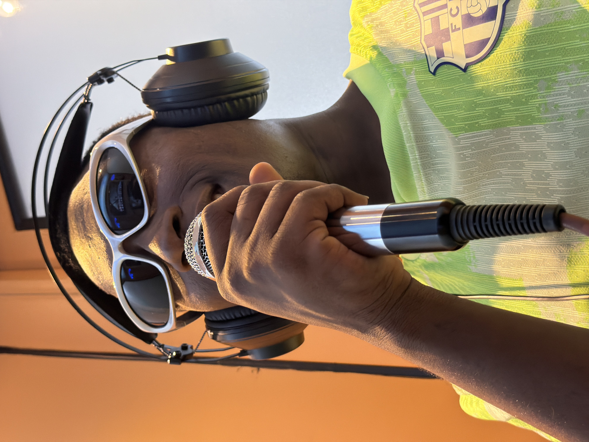 An individual is captured in a close-up, portrait-style shot, rotated 90 degrees clockwise from its original orientation, showing them engaged in what appears to be a recording or performance session. The person, with dark skin, is seen from the chest up, facing slightly away from the camera while holding a microphone to their mouth.
They are wearing a light green and white patterned short-sleeved shirt, which features a crest on the right side of the chest. The crest is primarily purple/blue and white, displaying the letters "FC" along with a stylized design resembling a football or emblem. White-framed, dark-lensed sunglasses cover their eyes, reflecting bright light sources or a screen. A pair of black over-ear headphones rests on top of their head, with wires visible running down.
The person's right hand grasps a silver and black microphone, with a mesh grille visible on the microphone head. Their left hand is also partially visible, supporting the microphone or resting near it. The background is simple, divided into a bright white or light-colored area behind their head and a warm, plain orange-brown surface that occupies the lower and right parts of the frame, possibly a wall or table. The overall setting appears to be indoors, possibly a studio or a room with soft, indirect lighting. The focus is entirely on the individual and their immediate activity. No specific location cues directly link the image to Jiangjin, China.