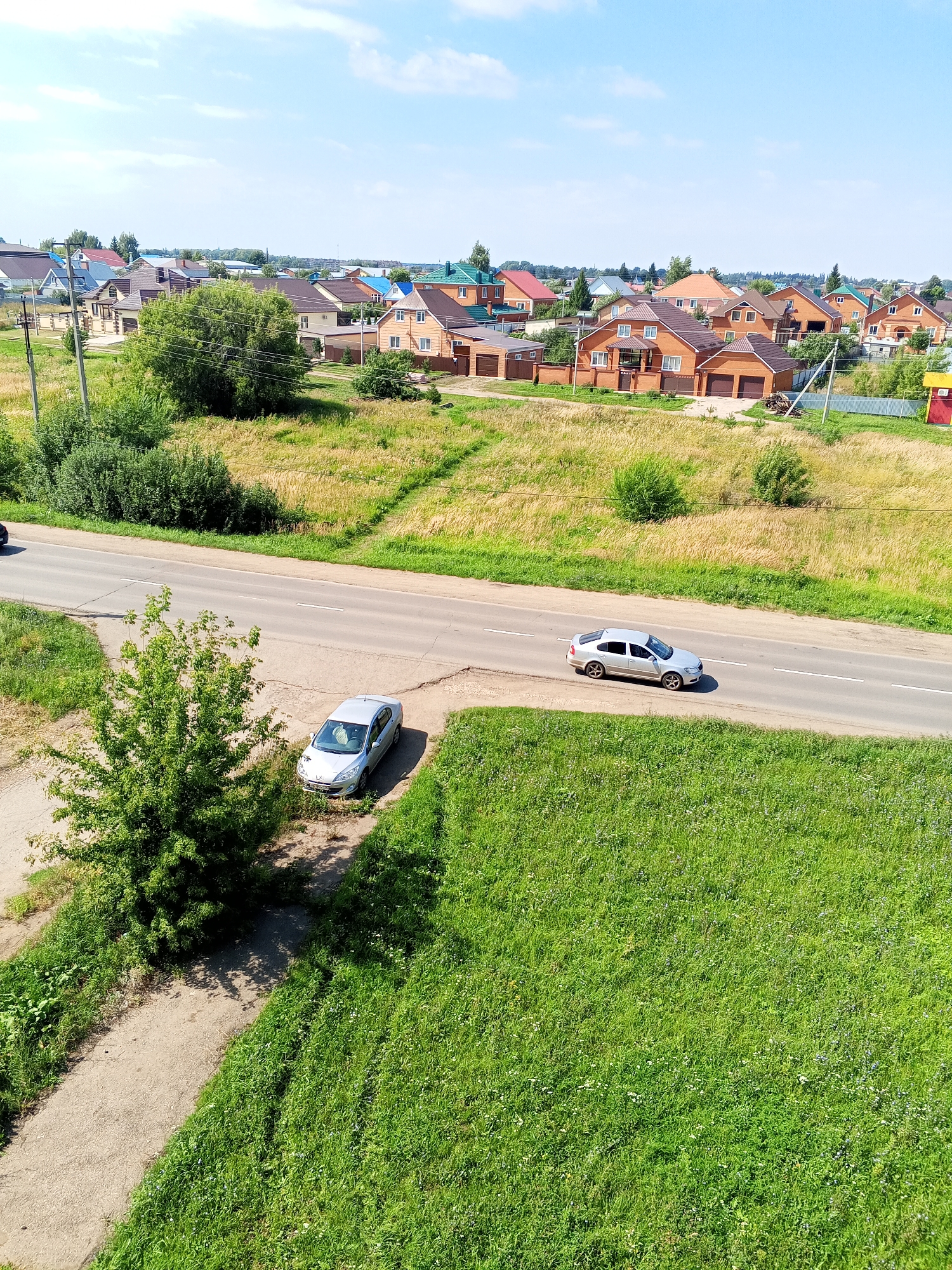A wide, overhead drone shot captures a rural residential area in Chistopol, Russia, on a sunny day. The scene features a two-lane asphalt road running horizontally across the middle of the frame. A silver sedan is driving on the right lane of the road, moving away from the viewer. Parked on the shoulder of the road on the left, a silver hatchback is visible.
To the left of the road, a patch of dry grass and dirt leads to a small, leafy green tree. Further left, a grey concrete path runs parallel to the road, bordered by lush green grass. The background showcases a cluster of residential houses with varied architectural styles and colored roofs, primarily oranges, browns, and blues. Some houses have attached garages, and fences delineate property lines. Tall green trees and bushes are scattered throughout the residential area. The sky above is clear blue with a few scattered white clouds.