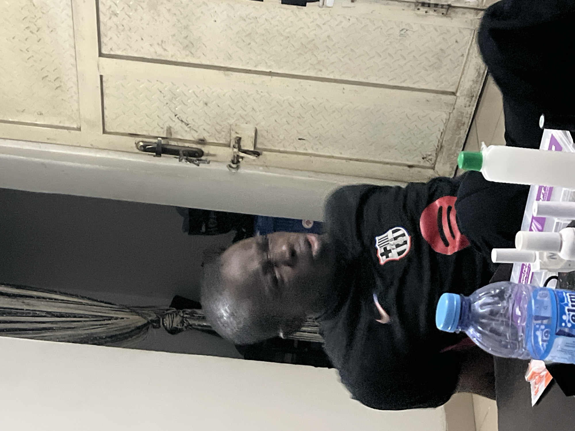 A man with a shaved head, wearing a black long-sleeved shirt with a white Nike logo and a red and black emblem on the chest, is sitting next to a table with a water bottle and medical supplies. He is in a room with a white door with a diamond plate design. It is not possible to definitively determine the location in the city of Abuja, Nigeria, without additional information.
