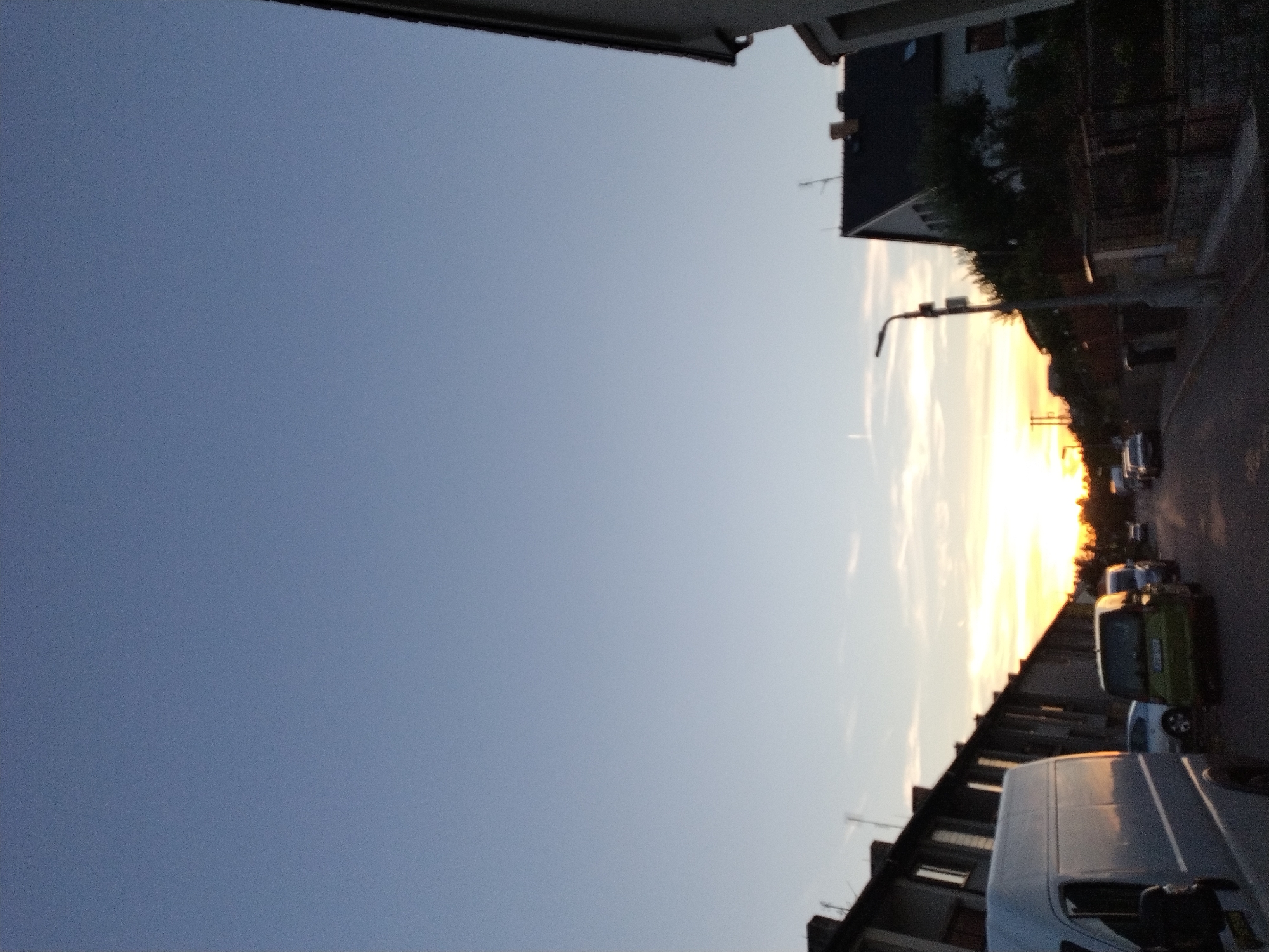 An exterior, eye-level, medium shot shows a residential street in Dobev, Czechia, during sunset. The sky is a pale blue with wispy clouds near the horizon, tinged with orange and yellow from the setting sun. The street is lined with houses and parked cars. In the foreground on the left is the side of a white van. Further down the street, there are several parked cars, including a green minivan in the middle of the road. A lamppost stands on the right side of the street. The overall lighting suggests dusk.