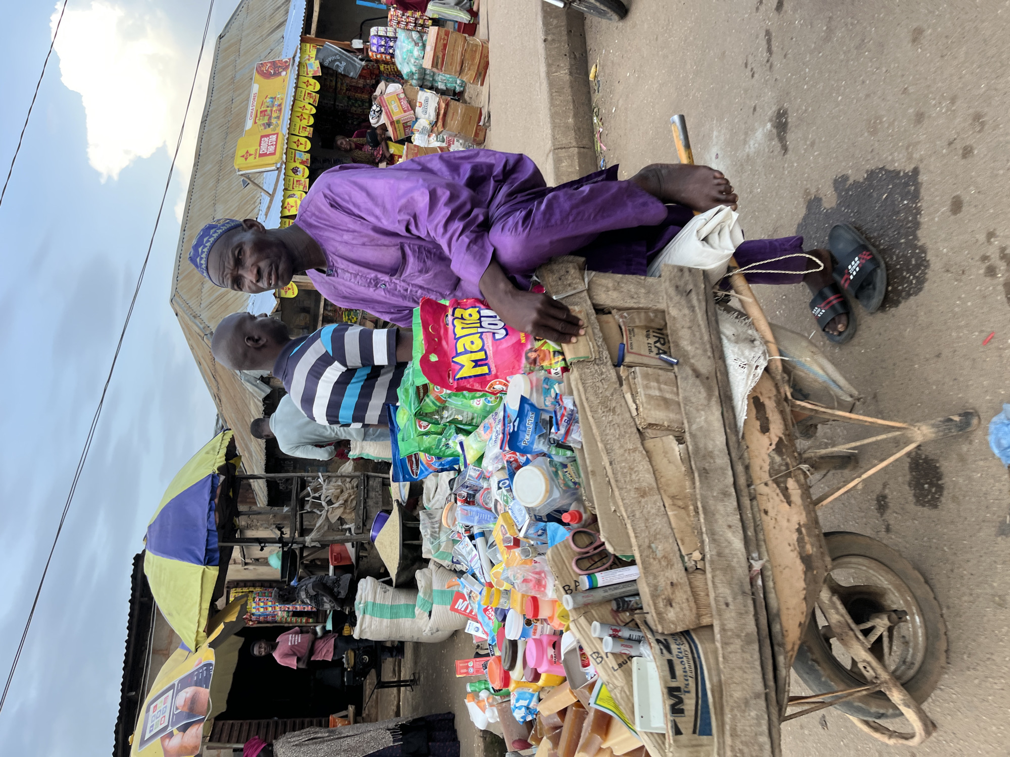 The image depicts a street market scene in Bukuru, Nigeria. In the foreground, a man wearing a purple traditional outfit and a blue cap is seated on a wooden cart filled with various goods, including packaged food items, bottles, and containers. He is looking towards the viewer. Behind him, several other people are visible, some interacting with market stalls. The stalls are covered with awnings, and displays of goods are arranged on shelves and on the ground. Bags of grain and sacks are also present. The environment appears to be an outdoor marketplace with a paved ground and a cloudy sky overhead.