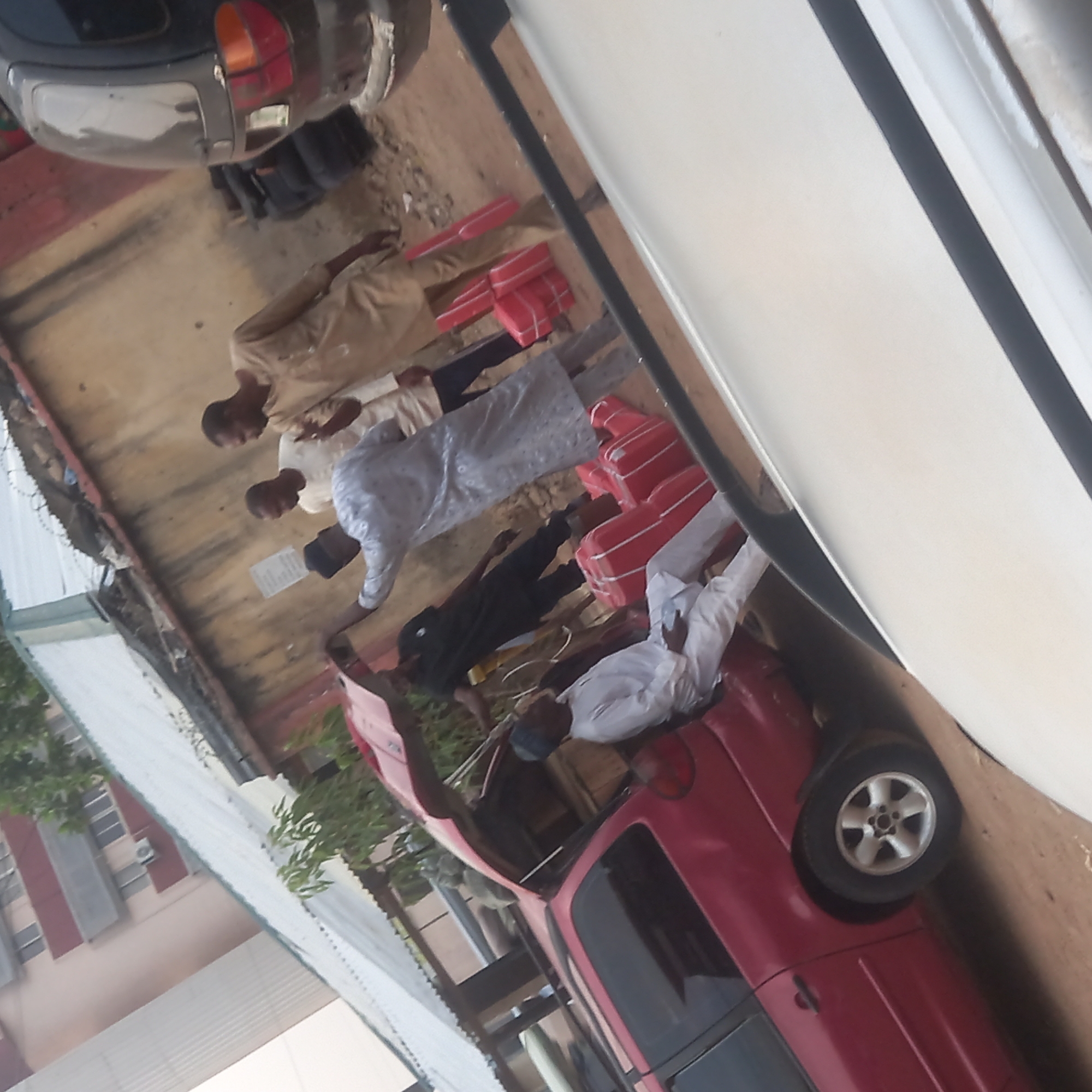 On a flat, concrete surface outdoors in Dadi Riba, Nigeria, several men are gathered around a red pickup truck and a white SUV. The men appear to be Muslim based on their attire. Several red packages are lined up between the vehicles and the men. Another dark-colored vehicle is partially visible in the background. Buildings are visible in the periphery of the image. The men are standing and talking amongst themselves.
