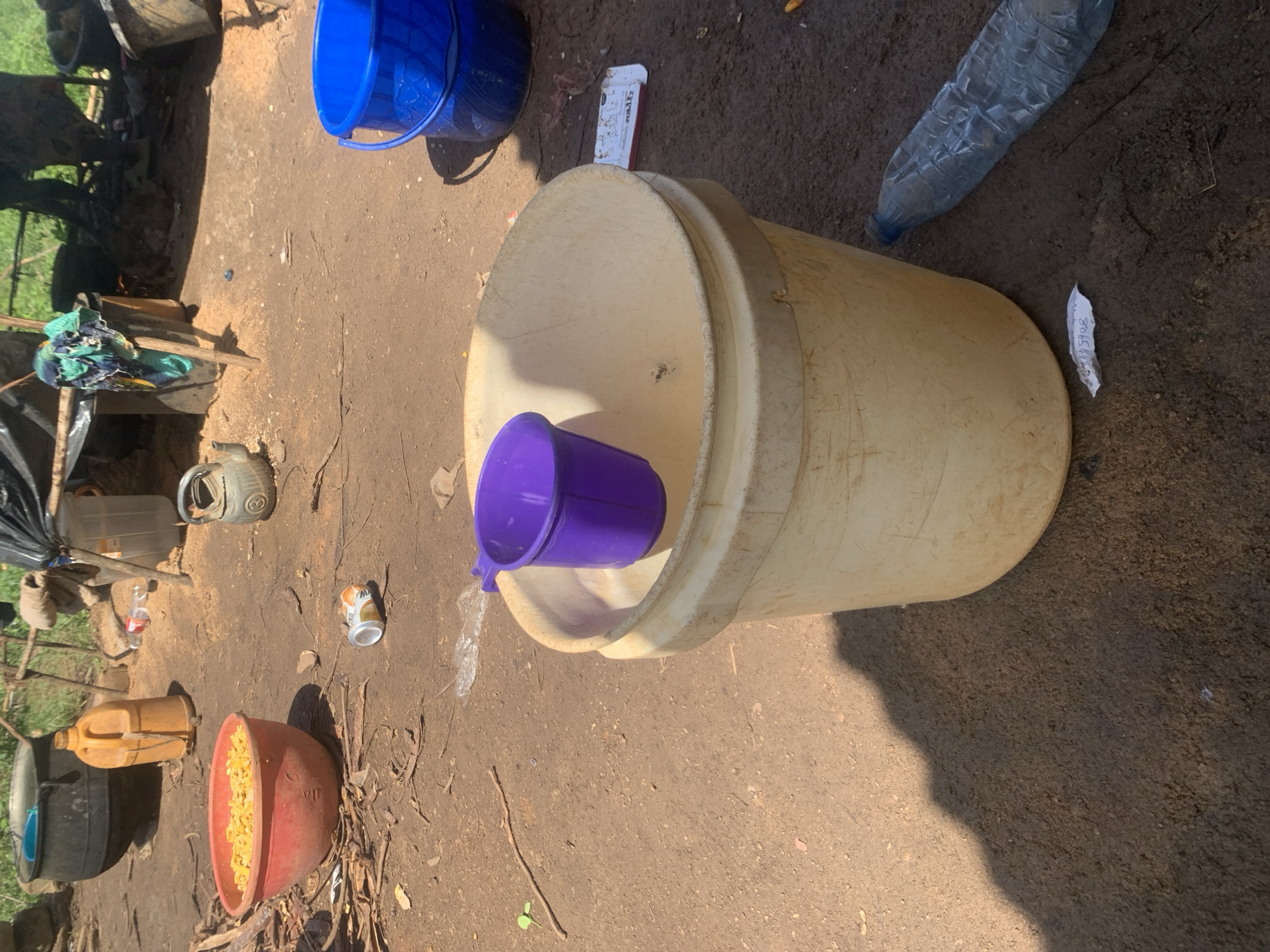This is a close-up, ground-level, eye-level shot of an outdoor scene in Buga da Gindi, Nigeria. The foreground is dominated by a large, tan plastic bucket containing a small, purple plastic cup. To the right of the bucket, a crumpled plastic water bottle lies on the dirt ground. Further back, a bright blue plastic bucket is visible. The ground is a mixture of dirt and wood shavings, suggesting an area of activity or construction. In the background, there are several large cooking pots, some with lids, and a collection of plastic containers and bottles. A makeshift shelter made of poles and tarpaulin covers some of the area. Scattered items on the ground include twigs, leaves, and what appears to be an empty aluminum can. The overall impression is of a functional, perhaps temporary, outdoor living or working space.