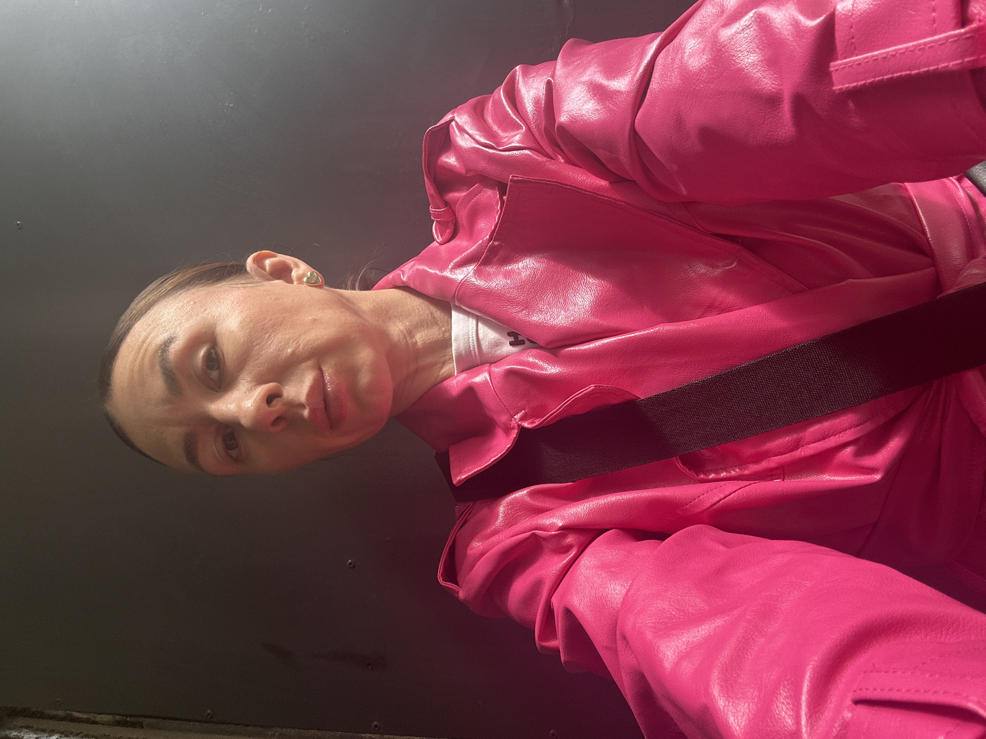 A woman with brown hair and eyes is wearing a bright pink jacket with a black strap across her torso. She is indoors against a dark gray wall. The jacket appears to be made of a glossy, possibly leather or vinyl-like material. Her facial expression is neutral. The image is taken from a low angle, looking slightly upward at her. The location, Votkinsk, Russia, cannot be determined from the visual information provided.