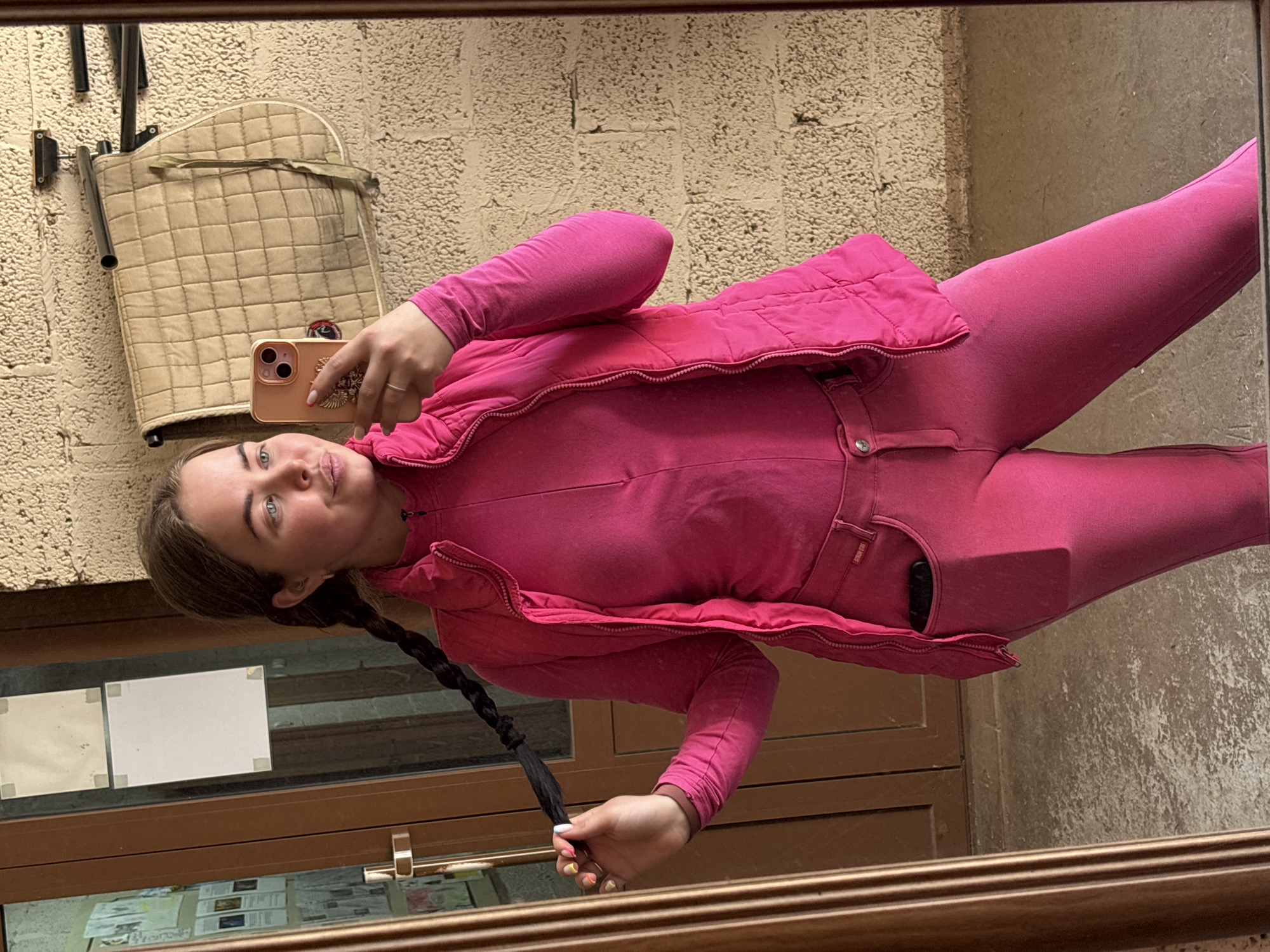 A young woman in a bright pink outfit is posing for a mirror selfie. She is wearing a matching long-sleeved shirt, pants, and an unzipped vest. Her hair is styled in a braid. She holds a smartphone in her right hand, taking the picture. In the background is a wall and a padded object hanging from it. A window or door is visible below the woman, with papers displayed behind the glass. Based on the information provided, the location is Русский Вожой, Russia.