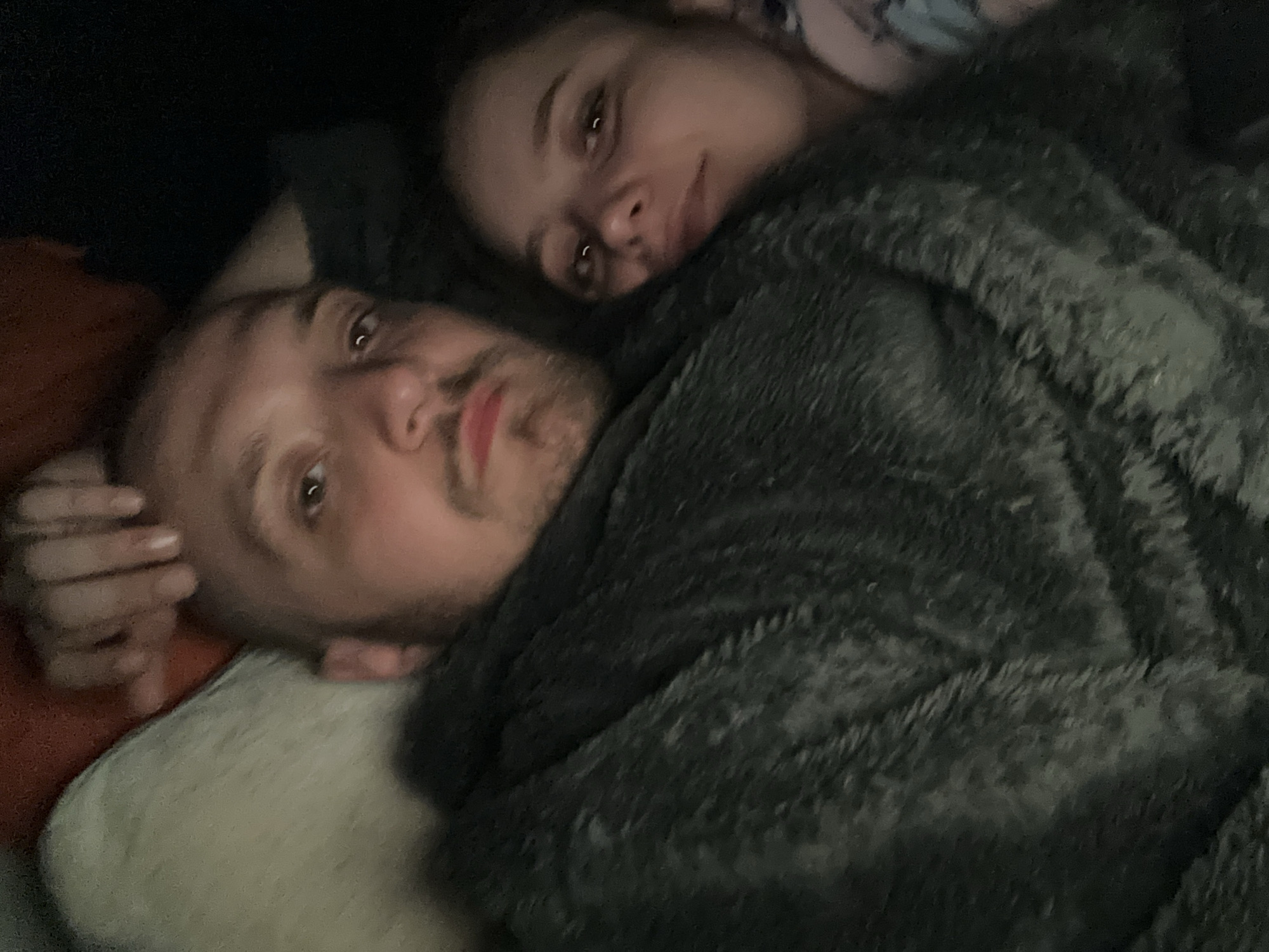 Two people are lying down under a gray blanket. One person, with lighter skin and short hair, is lying on their back on a pillow. Their left hand is visible near their head. The other person is lying on their side, closer to the camera, with their head resting near the first person. The environment is dimly lit. The city location is not discernible from the image.