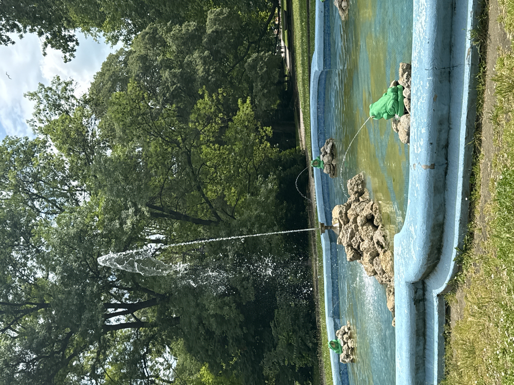 A bright blue, tiered fountain with a central rock formation and water jets is positioned in a park. Several green, cartoonish frog statues are placed around the fountain's edge, some of which are acting as water spouts. A tall, thin water jet erupts from the center of the fountain. Lush green trees and a cloudy sky form the backdrop. The ground in front of the fountain is covered in grass and small white flowers. In the distance, a paved path winds through the park, with a few people visible walking along it. The scene appears to be in a public park on a sunny day.