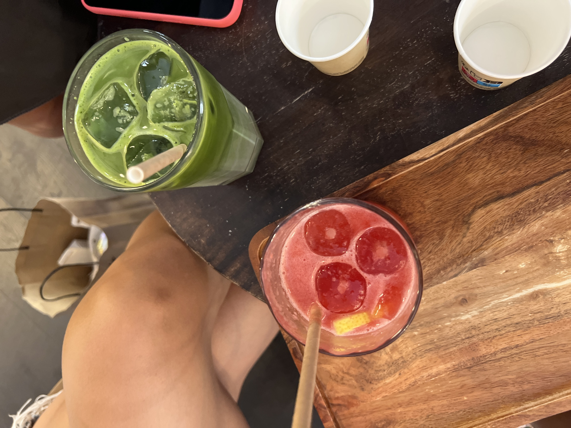 A close-up, overhead shot shows two drinks on a dark wooden table, along with a paper bag and part of a person's leg. In the foreground, on the left, is a glass of red liquid with three red ice cubes and a slice of yellow citrus fruit. A light-brown straw extends from the drink. This glass sits on a wooden tray. To the right, in the background, is a tall glass of vibrant green liquid filled with ice cubes. A light-colored straw is visible in this drink. Also on the table are two empty paper cups. Partially visible in the upper left of the image is a person's bare leg and thigh, with a white garment draped over it. Behind the drinks, a brown paper bag is partially visible. The image does not contain any specific landmarks or features that would allow for the identification of a location in Paris, France.