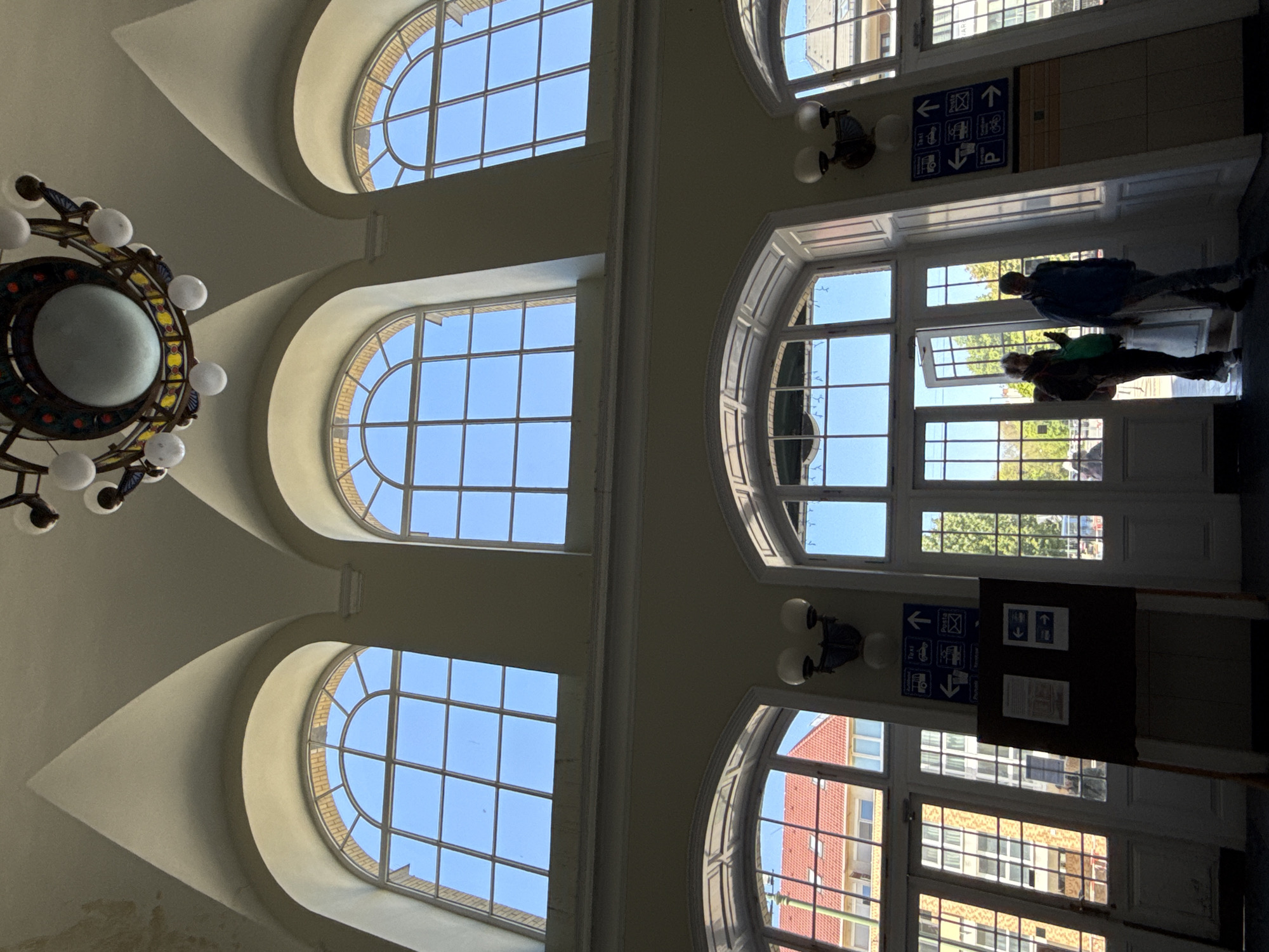 This media file captures the interior of a grand, high-ceilinged building, identified as a transport hub, likely a train or bus station in Szeged, Hungary. The image is oriented vertically, showing the ceiling and upper walls on the left, and a wall with windows and a doorway on the right.
The architecture is notable for its arched and curved ceilings and numerous large windows. On the left side of the image (which would be the ceiling if correctly rotated), three large, semi-circular arched windows illuminate the space with natural light, showcasing a clear blue sky. A large, ornate chandelier with multiple white globe lights hangs prominently from the ceiling.
On the right side of the image (a wall), a series of rectangular and arched windows, along with glass doors, provides further illumination and views of the exterior. Through these windows, a bright, clear blue sky, parts of other buildings (including red brick and yellow/beige structures), and green trees are visible, indicating a sunny daytime setting.
Two individuals are silhouetted against the bright light from the doorway on the right, appearing to walk or stand near an entrance/exit. One person wears a blue jacket and dark pants, while the other wears a darker top, lighter pants, and appears to carry a green bag.
Mounted on the wall on the right is a blue informational sign with white pictograms and Hungarian text. Visible text includes "Post", "Taxi", "Poggyász" (luggage, likely indicating a train station), "Autóbusz" (bus), "Kerékpár" (bicycle), and "Parkolás" (parking), accompanied by directional arrows. Below this, a dark board contains additional, partially obscured information. Two smaller, wall-mounted light fixtures with two globe lights each are also visible on this wall.