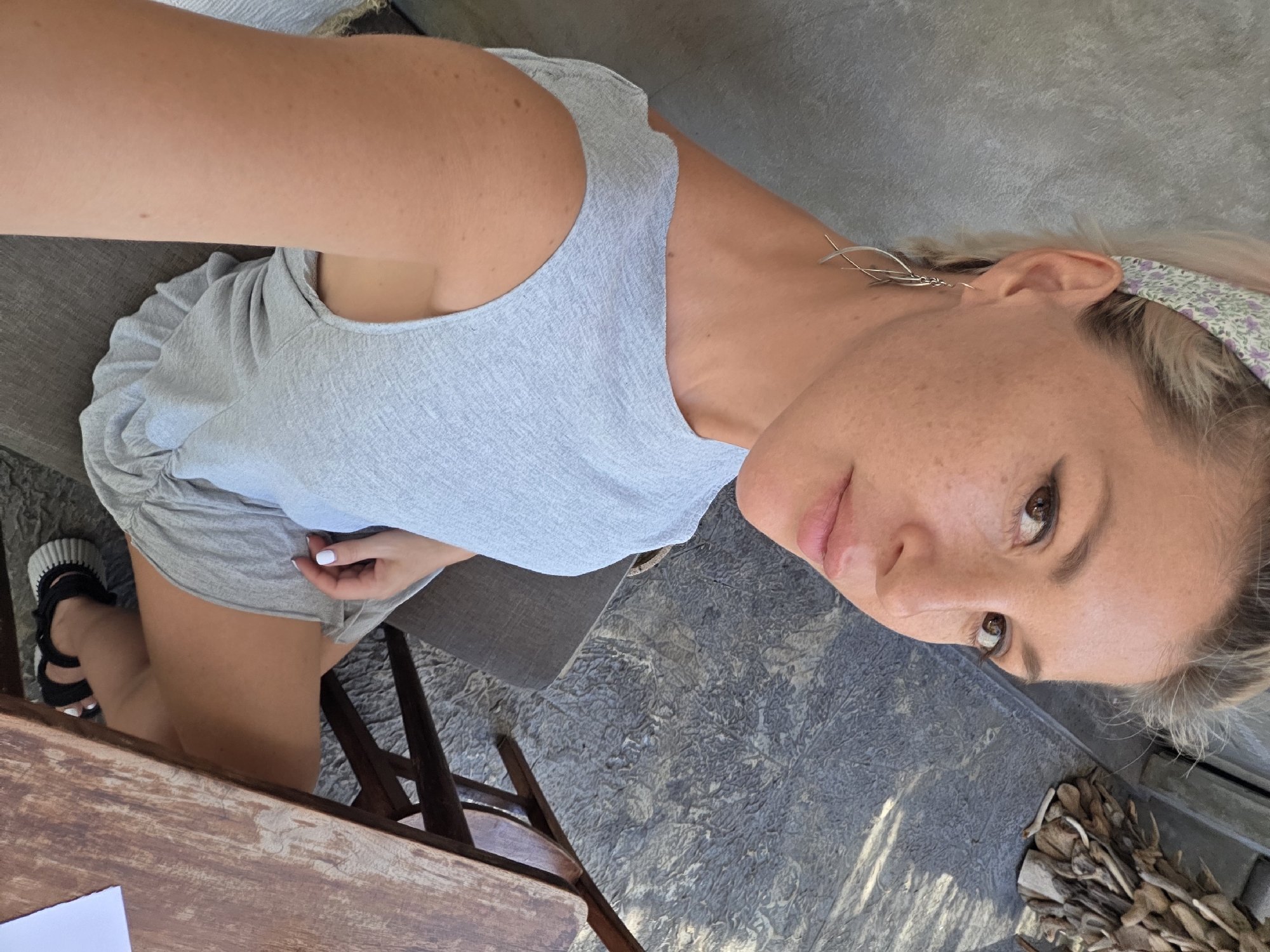 A woman with fair skin, freckles, and light blonde hair is sitting at a wooden table. She wears a light blue-gray dress, black sandals with platform soles, and a floral headband. She has silver earrings in her left ear. Her fingernails are painted white. The woman is looking at the camera and is situated on a patio or outdoor seating area, with a gray textured floor. There is a pile of sticks or kindling in the lower right corner. The location is consistent with a residential or commercial outdoor space in North Kuta, Indonesia.
