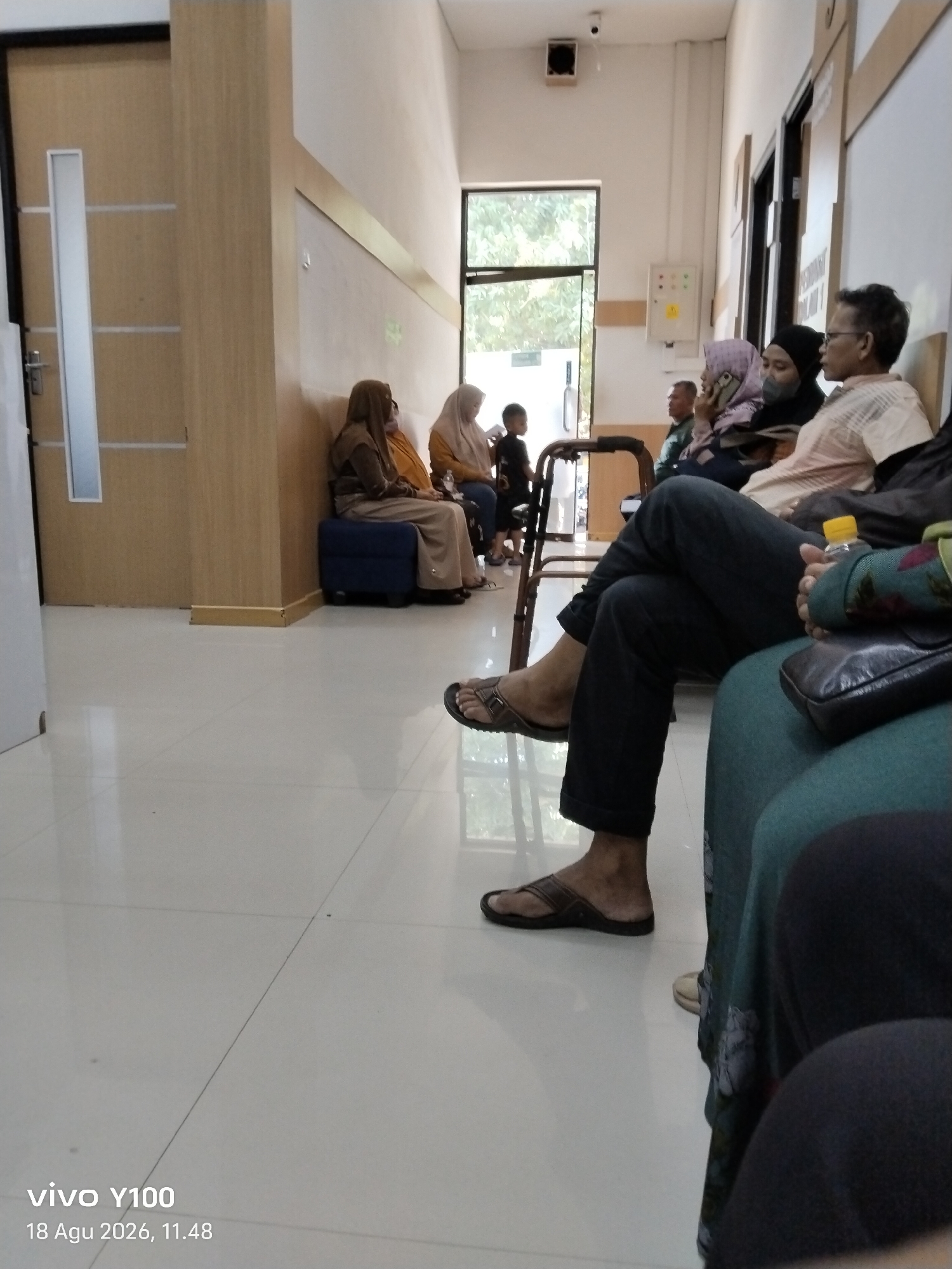The image depicts an indoor waiting area, possibly a clinic or government office, in Patokan, Indonesia. Several individuals are seated on a long bench and chairs. In the foreground, a person's legs and feet are visible, wearing dark pants and brown flip-flops. To the left, a wooden door with vertical white panels is visible, with a metal handle. The walls are light-colored, with a wooden trim along the bottom. A large window with a view of greenery is present in the background. The floor is tiled and appears to be white or light grey. The timestamp at the bottom indicates "vivo Y100" and "18 Agu 2026, 11.48".