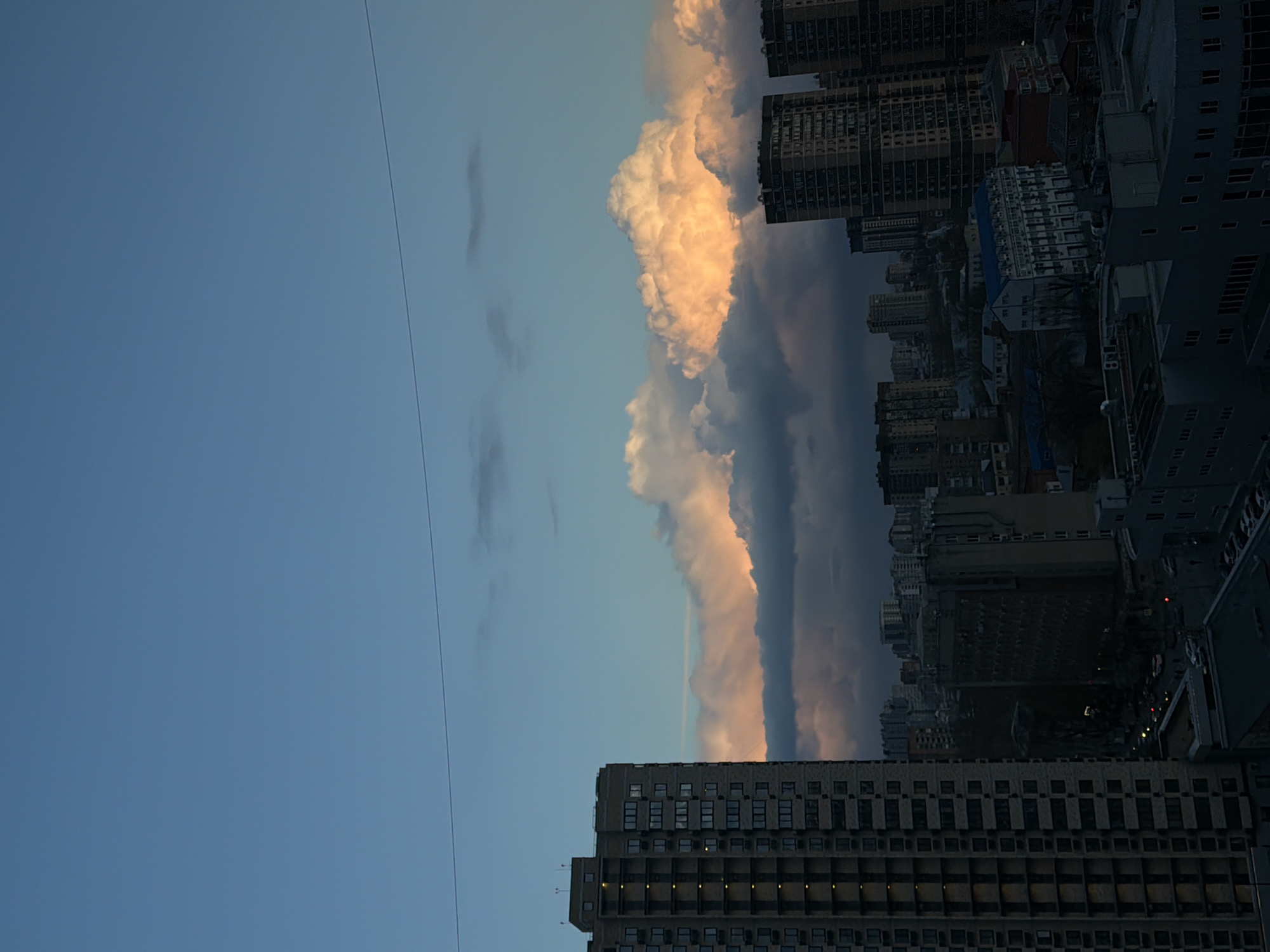 The image depicts a cityscape with buildings silhouetted against a sky dominated by dramatic cloud formations. The scene is likely captured from a high vantage point within Rostov-on-Don, Russia. The sky transitions from a clear blue to a band of thick clouds that are illuminated with a soft, warm light at the top, contrasting with the darker, more ominous clouds below. These clouds create a distinct vertical band in the center of the image. Buildings, predominantly high-rise apartments and structures, occupy the right side of the frame and the bottom edge. A power line cuts diagonally across the upper left portion of the sky. No people are discernible. The image's overall tone suggests a contrast between urban density and the transient, atmospheric beauty of the sky.