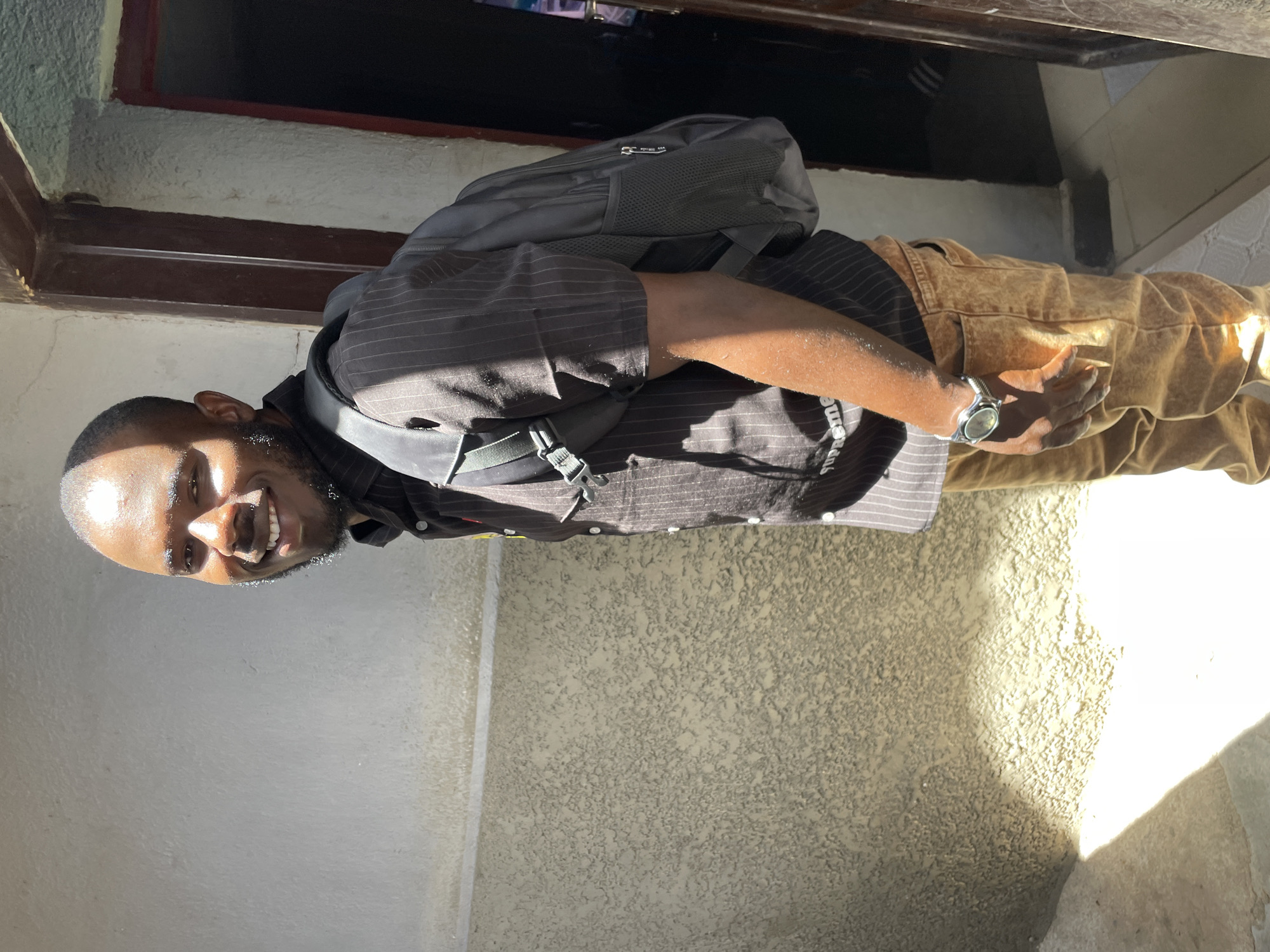The image shows a Black man, likely in Jos, Nigeria, standing in what appears to be a brightly lit doorway or passage. The man is smiling broadly and looking directly at the camera, conveying a friendly and approachable demeanor. He has a shaved head and a short beard.
He is dressed in a dark, possibly black or dark grey, short-sleeved collared shirt with thin vertical stripes. A grey backpack is worn over both shoulders. On his left wrist, he wears a silver-toned watch. He is wearing light brown or khaki-colored cargo pants. Partially visible text on his shirt, near his left arm, reads "Supreme".
The setting appears to be an indoor or semi-outdoor space, characterized by textured walls and ground in shades of light beige and grey, resembling stucco or concrete. A dark rectangular opening, possibly a doorway or window, framed by dark wood, is visible in the upper background. Strong, direct sunlight illuminates the scene, casting harsh shadows on the man's right side and on the textured surfaces, suggesting it is daytime.