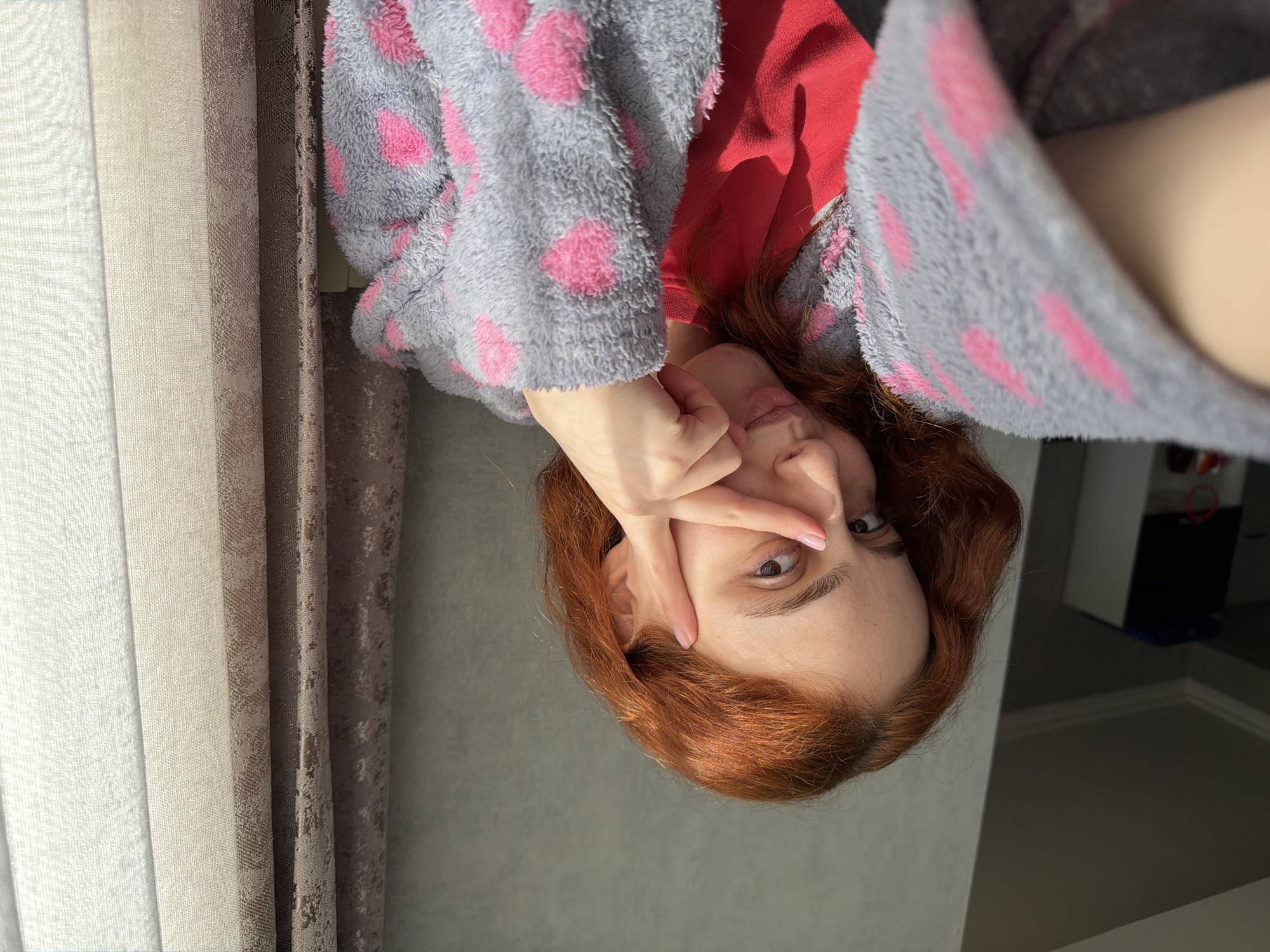 A woman with auburn hair is photographed indoors. She wears a gray robe with pink hearts over a red top and has her hand raised near her face. She is in front of curtains of beige, white, and brown. The location cannot be determined as Tbilisi, Georgia, without additional information within the image.