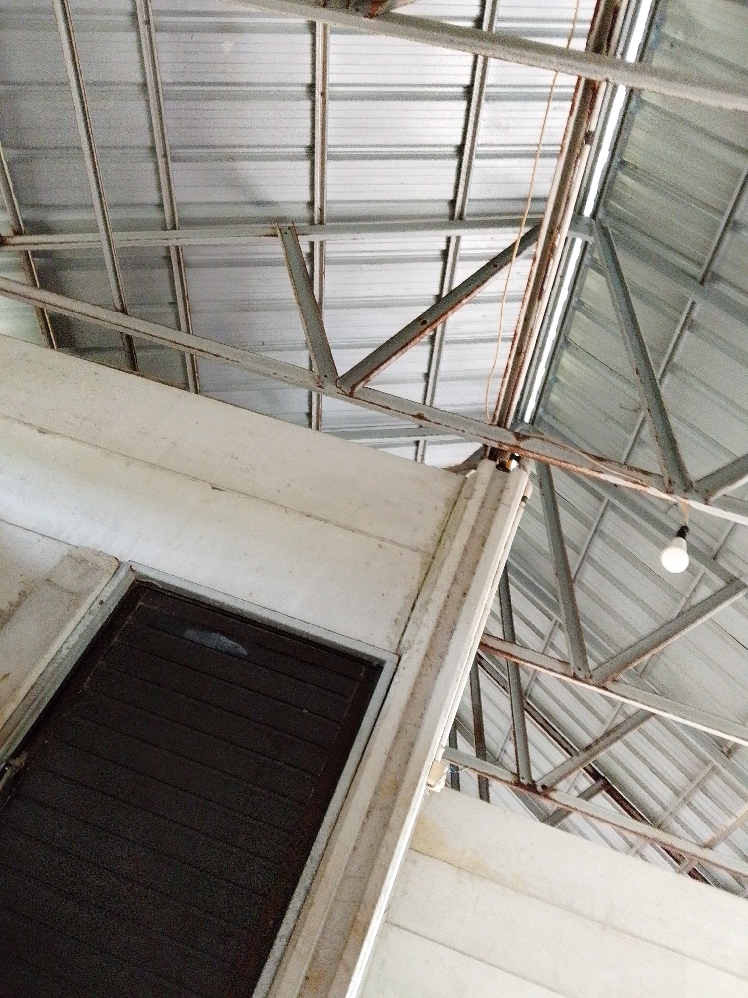 A low-angle shot of the interior structure of a building in Bukit Ketok, Indonesia, reveals a corrugated metal roof supported by a network of steel beams. The roof's metal sheets run in parallel lines, with the beams forming triangular and linear supports. A single light bulb hangs from the steel framework. Adjacent to the roof structure, a white painted wall with a dark brown, shuttered window or door is visible. The building appears aged with slight rust and discoloration of the metal and wall.