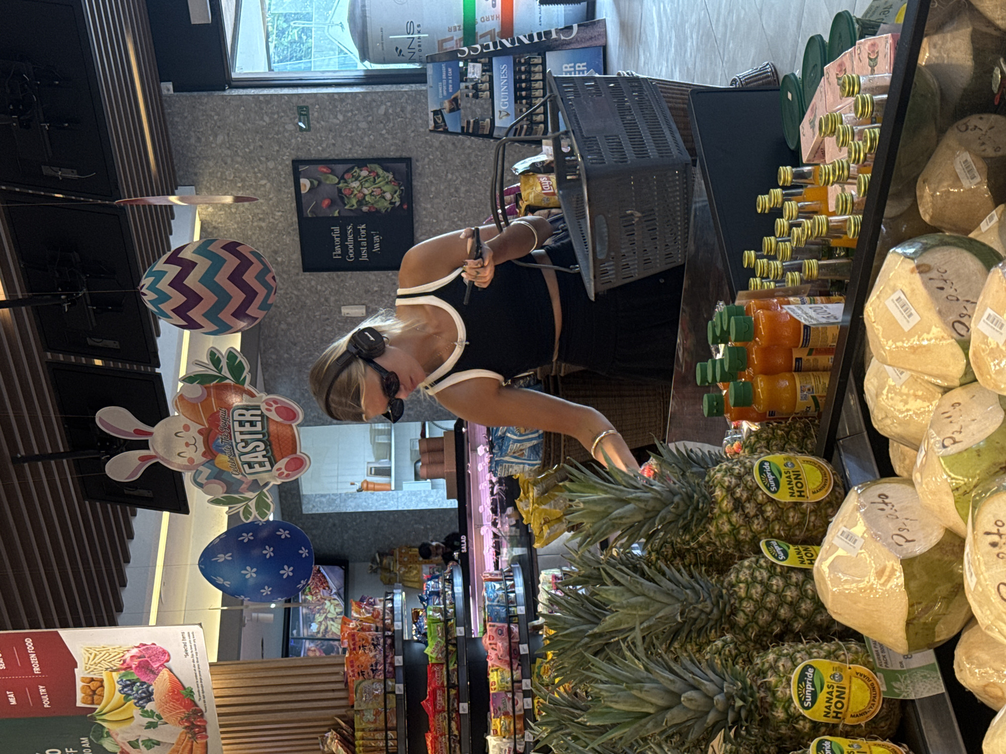 The image shows a woman in a black and white top, wearing sunglasses and headphones, shopping in a supermarket. She is reaching for a pineapple. The supermarket aisles are filled with various products. Easter decorations hang from the ceiling. There are coconuts and bottles of juice on the shelf beside her. Products from the brand Guinness are visible in the background. The pineapples she is reaching for are branded "Sunpride Nanas Honi." The scene appears to be a supermarket in North Kuta, Indonesia.