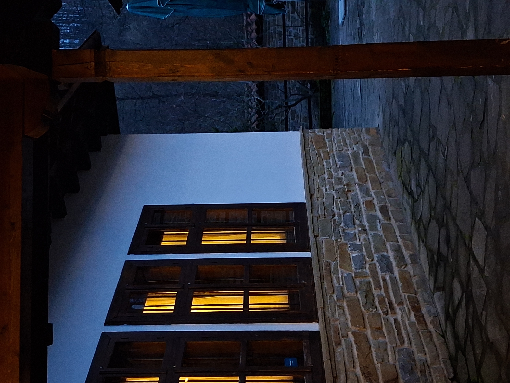 Captured outdoors in what is likely Katunishte, Bulgaria, the image presents an architectural scene at twilight or night. The focal point is a building exterior marked by a white-painted wall adorned with windows. Each window glows with light, suggesting occupancy or interior illumination. To the right of the wall is a constructed brick structure. An overhanging beam suggests a porch or roof extension. The ground is paved with stone tiles. Background foliage, obscured by darkness, hints at surrounding trees or vegetation.