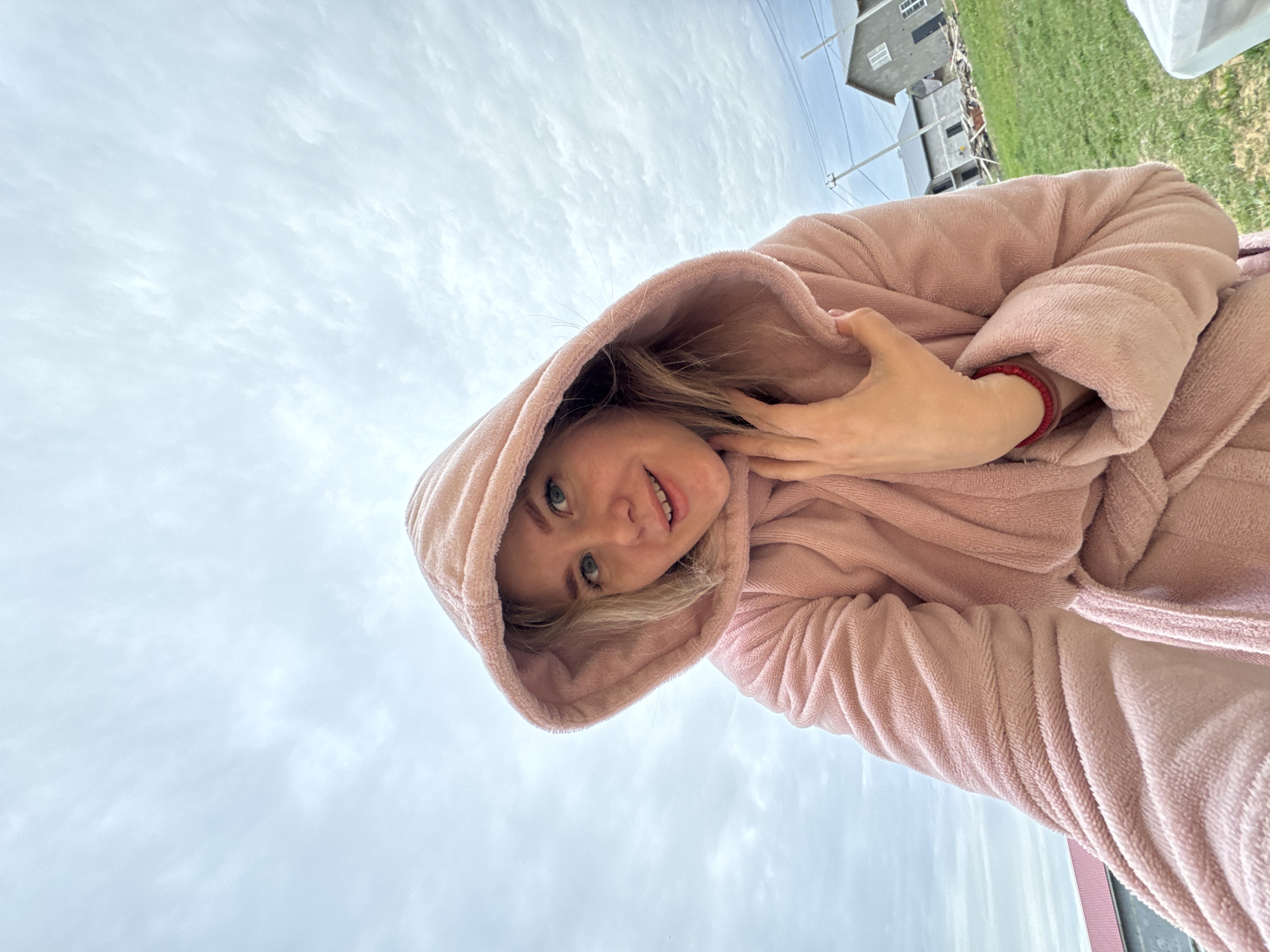 A woman in a pink hooded robe looks down at the camera. She has fair skin, blonde hair, and blue eyes, and her left hand is at her neck. The background shows a cloudy sky and a building with green grass. The location is Ferma, Russia.